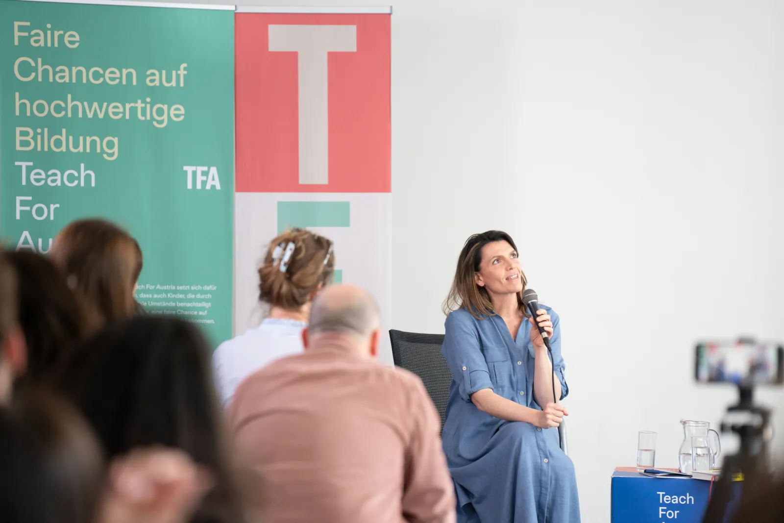 Person sitzt auf Stuhl, spricht ins Mikrofon; Publikum im Vordergrund; linkes grünes Banner mit 'Faire Chancen auf hochwertige Bildung'.
