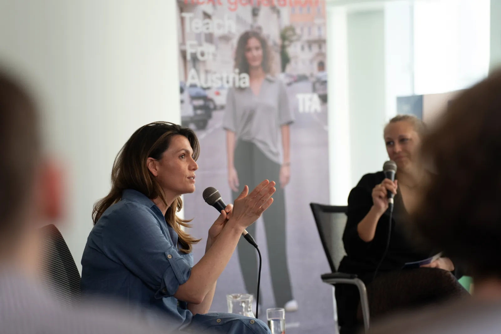 Sprechende Person bei Paneldiskussion spricht ins Mikrofon; daneben hält eine weitere Person ein Mikrofon, Banner im Hintergrund.