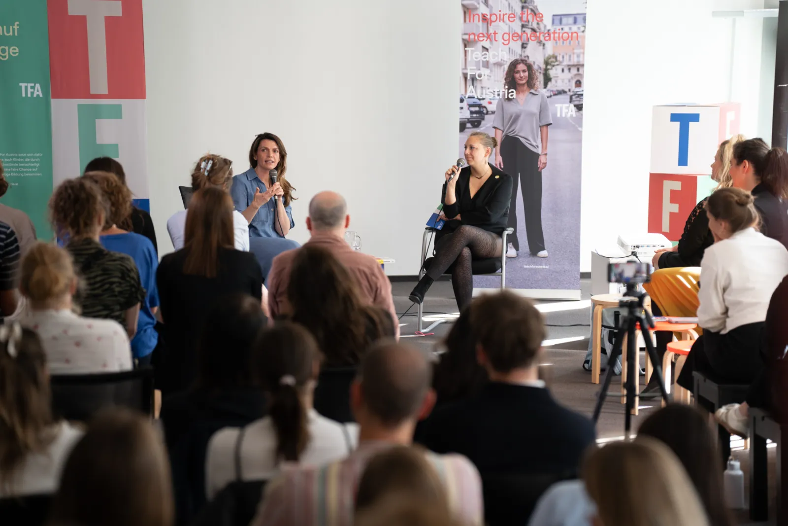 Zwei Moderatorinnen führen Podiumsdiskussion vor Publikum; Mikrofone, Banner im Hintergrund, Kamera auf Stativ im Raum.