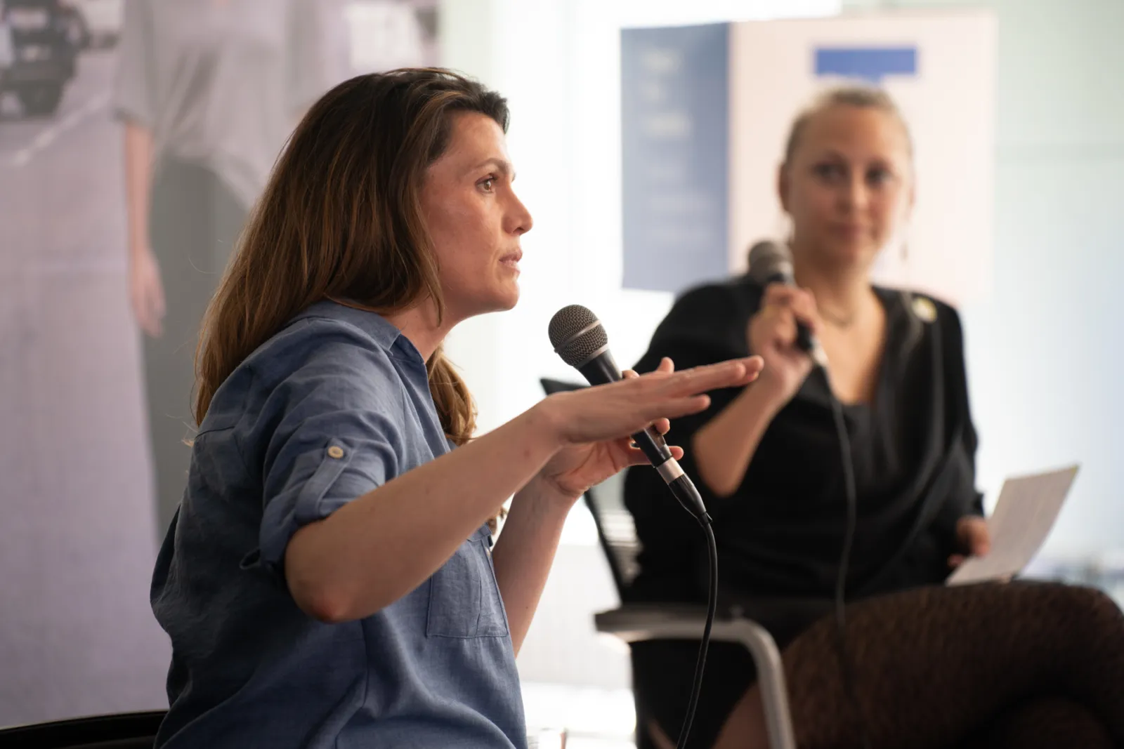 Person in blauem Hemd spricht ins Mikrofon und gestikuliert; im Hintergrund sitzt eine weitere Person mit Mikrofon bei Diskussionsrunde.