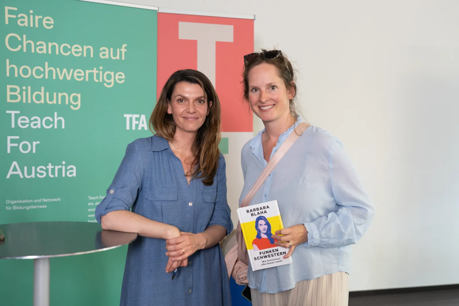 Zwei Personen stehen lächelnd vor Bannern von Teach For Austria; eine Person hält Buch mit Barbara Blaha.