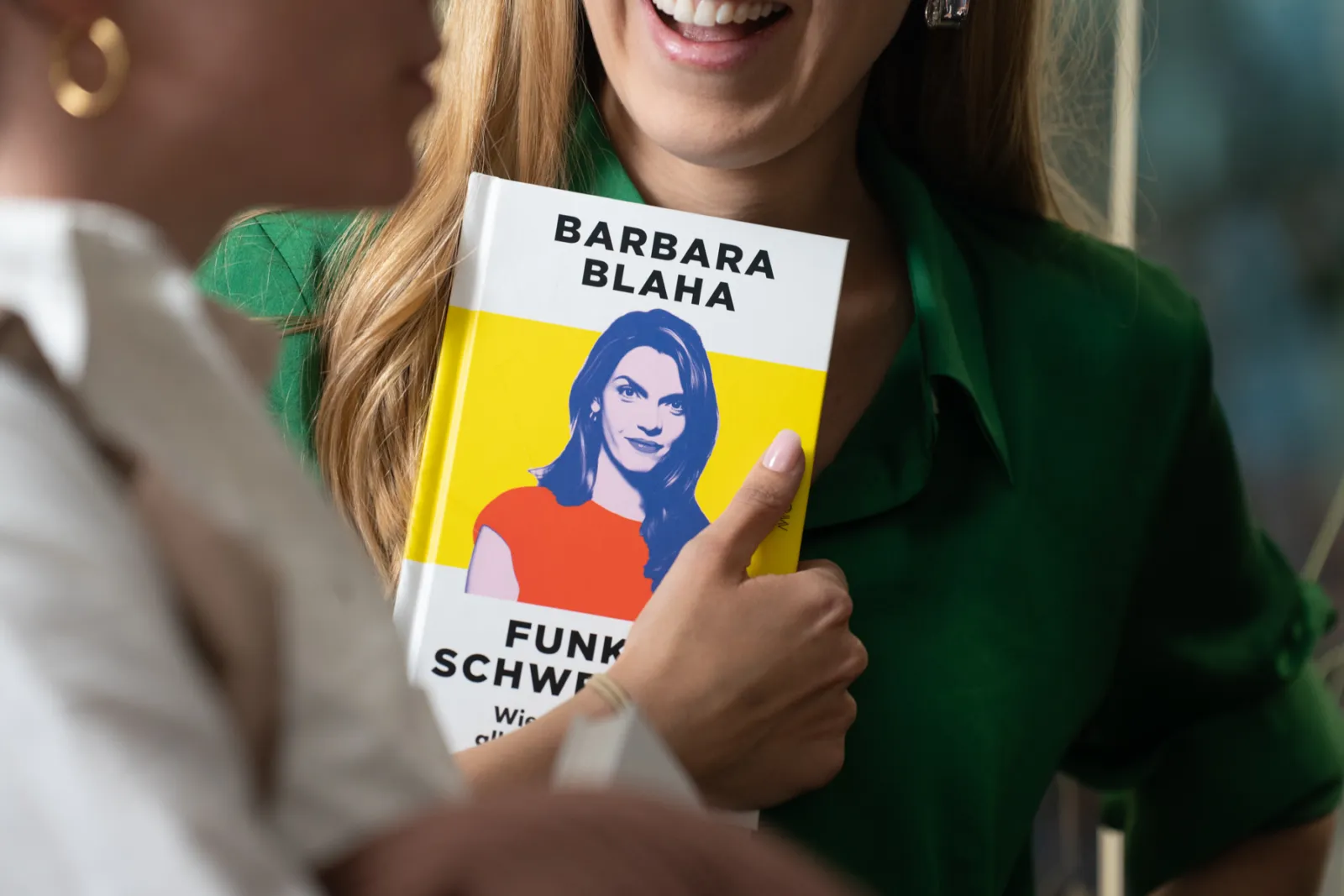 Eine Person hält ein farbiges Buch mit Barbara Blaha-Cover vor der Brust; Nahaufnahme, grüne Bluse.