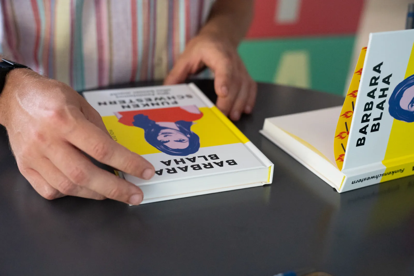 Person legt farbiges Buch auf einem Tisch, weitere Bücher stehen daneben.