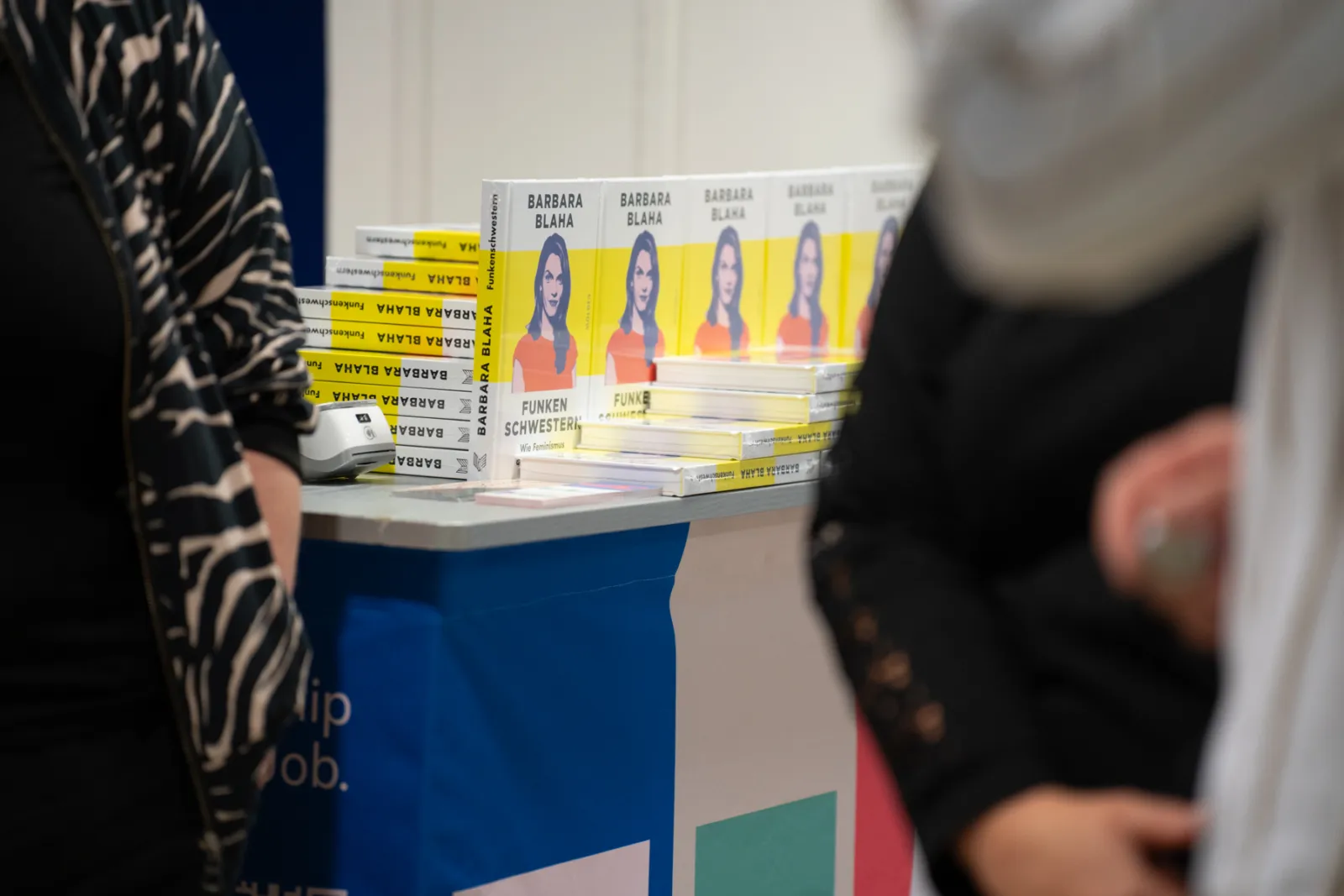 Bücherstand mit gelben Barbara Blaha-Coverbüchern; gestapelte Exemplare, Passanten betrachten das Display.