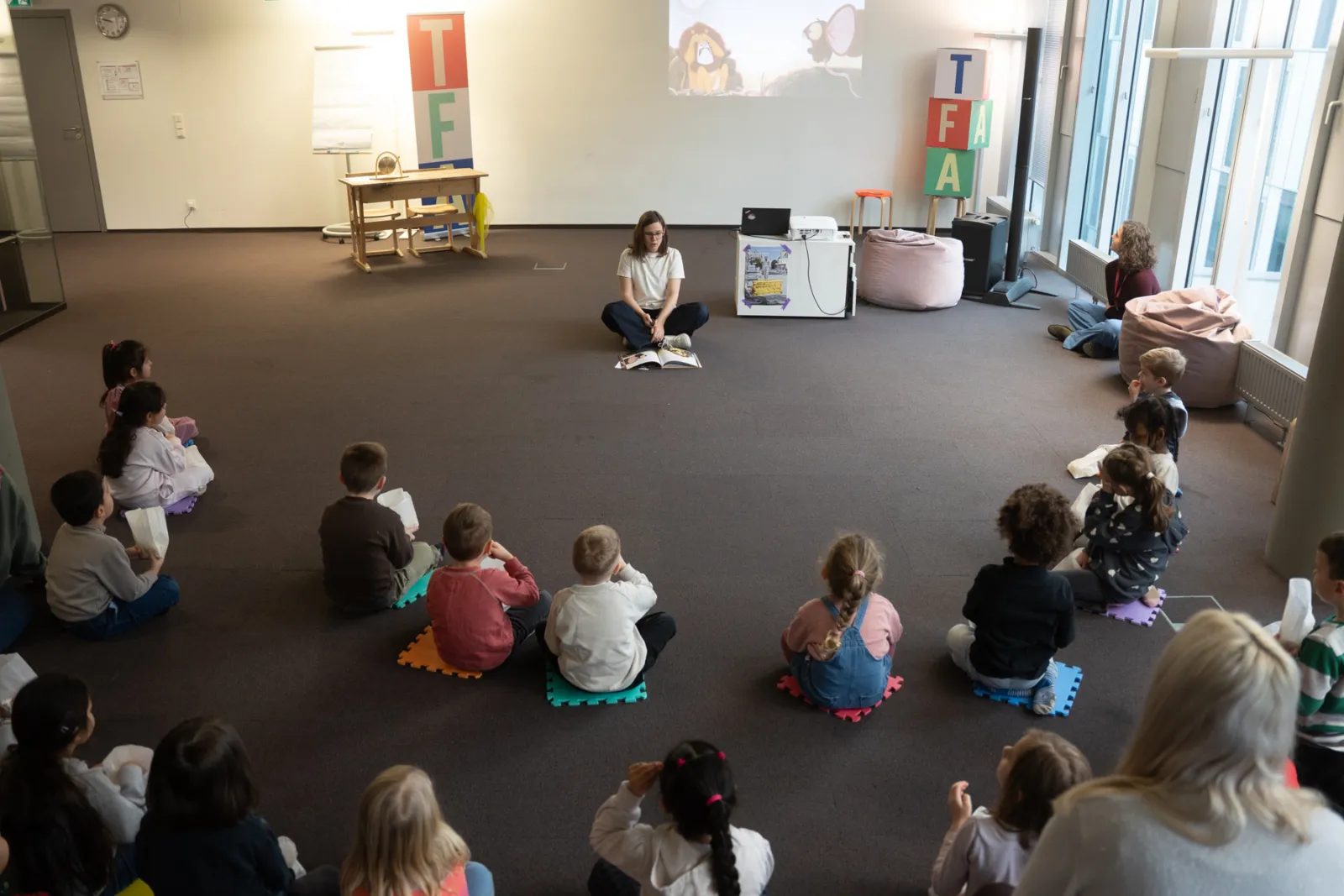 Lehrperson liest Kindern aus einem Buch vor; Gruppe sitzt kreisförmig auf bunten Schaumstofffliesen in einem Leseraum.