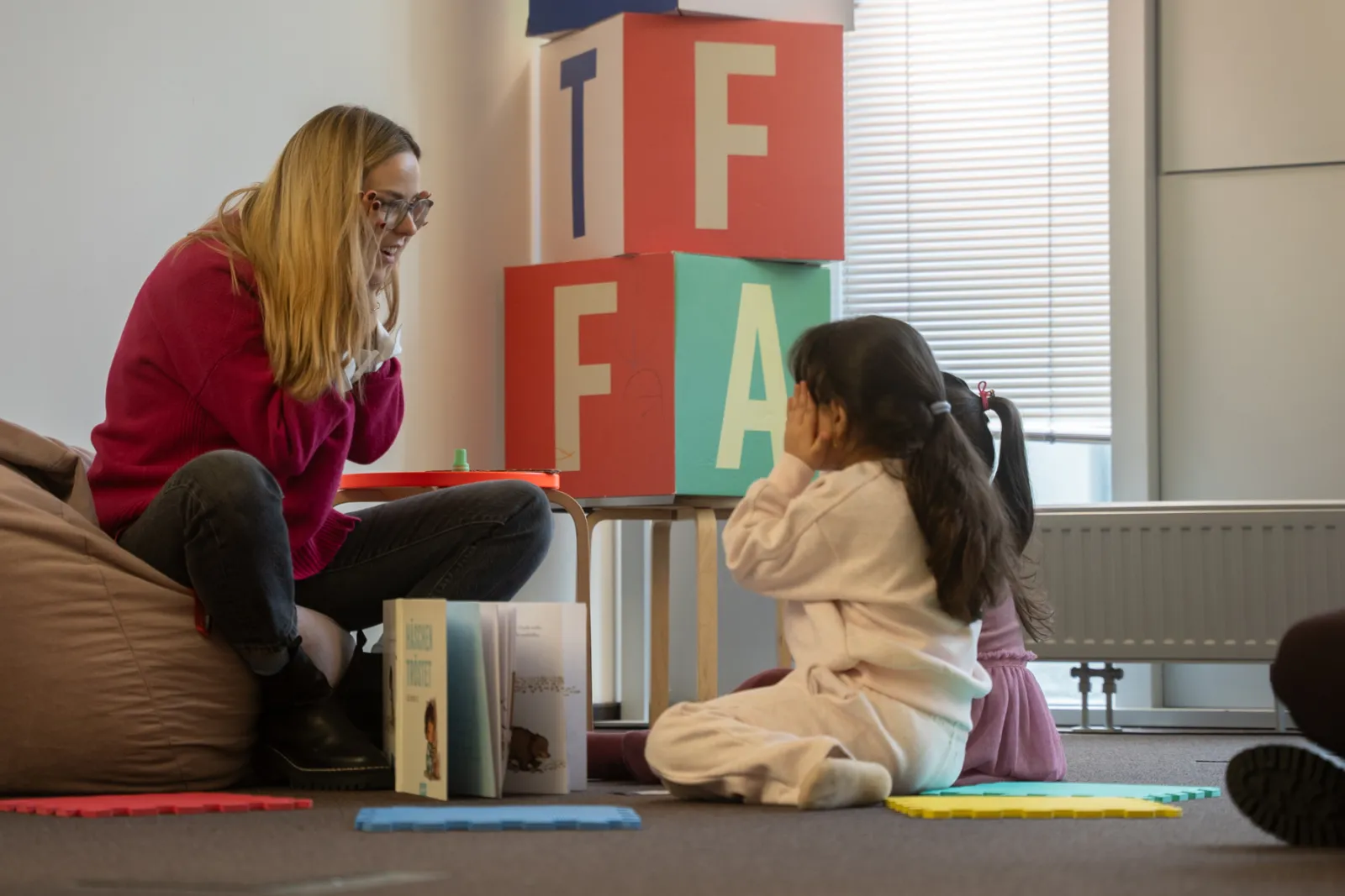 Erwachsene Person sitzt auf einem Sitzsack und spricht mit Kind auf dem Boden; bunte Spielbausteine und Bücher im Spielzimmer.