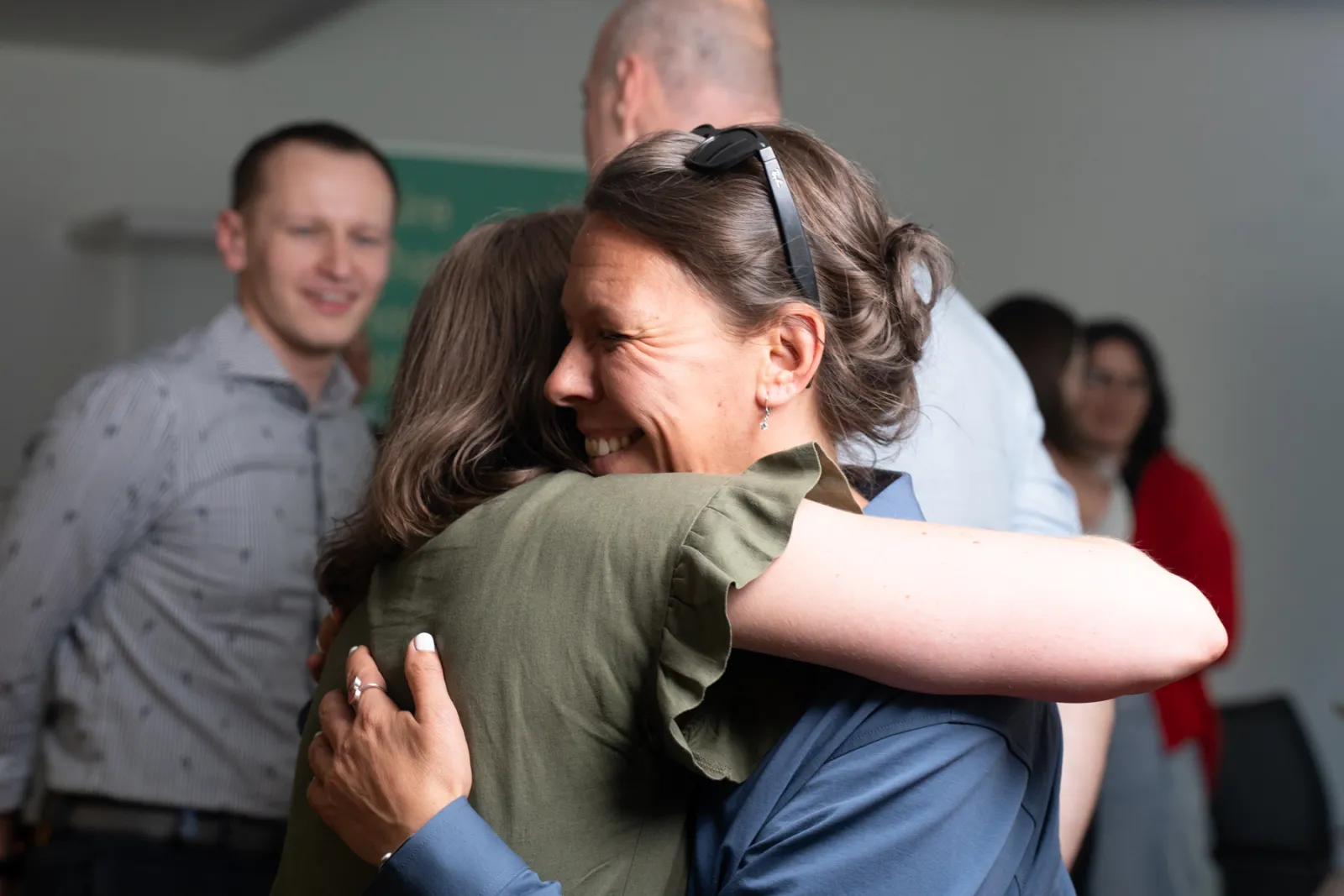 Zwei Erwachsene umarmen sich in einem Innenraum; im Hintergrund stehen weitere Personen.