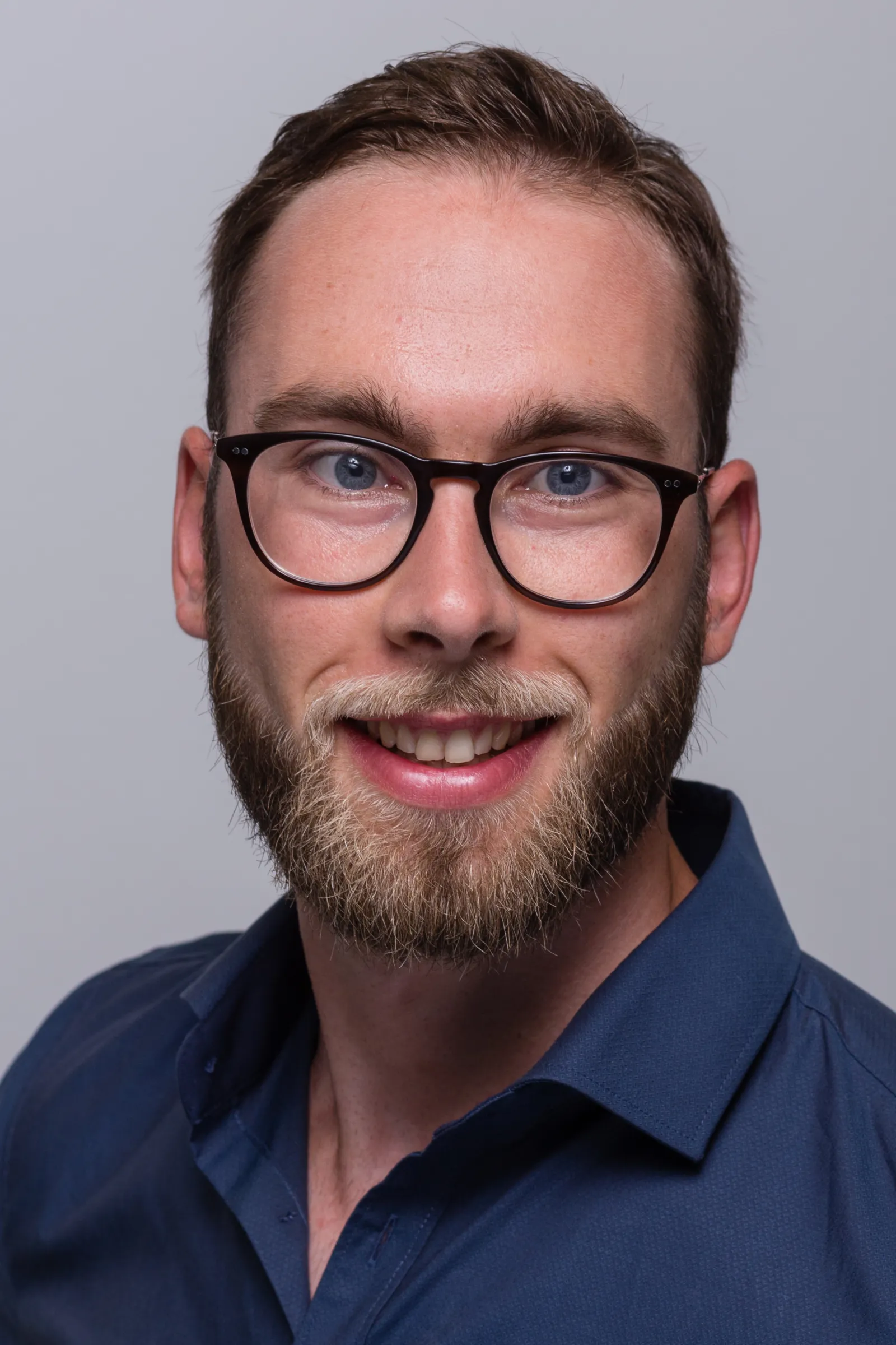 Mann mit Brille und Bart lächelt in die Kamera; Studio-Porträt vor grauem Hintergrund.