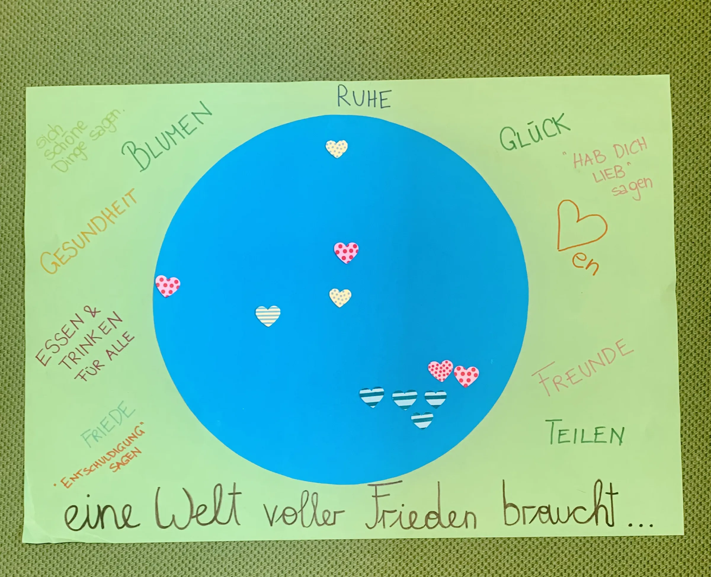 Blaue Kreisscheibe auf hellgrünem Hintergrund; Herzen und Text am Rand: Ruhe, Glück, Freunde, Teilen; unten: eine Welt voller Frieden braucht