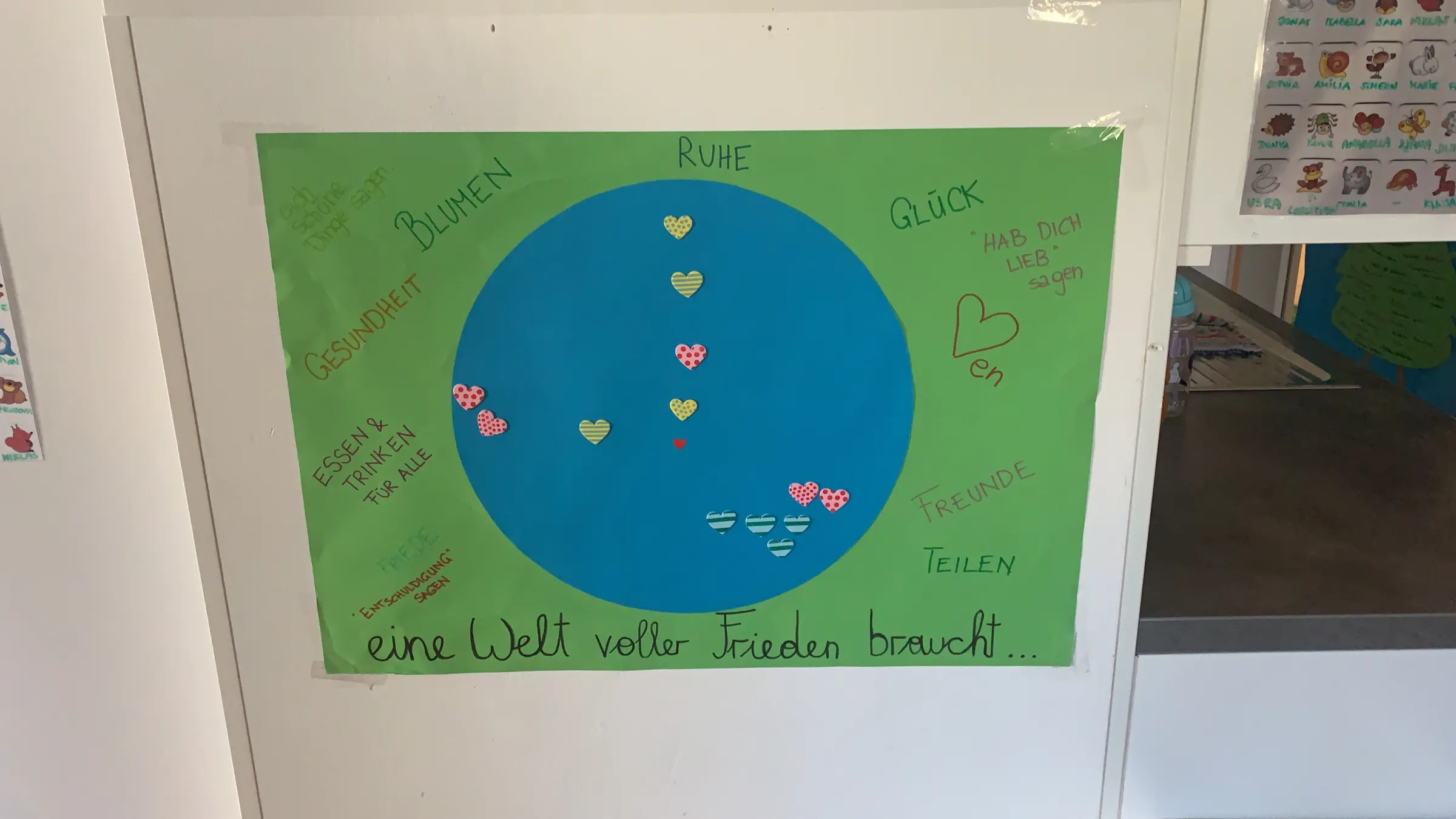 Plakat mit grünem Hintergrund, blauem Kreis als Erde, Herzsymbole; Text: 'eine Welt voller Frieden braucht'.