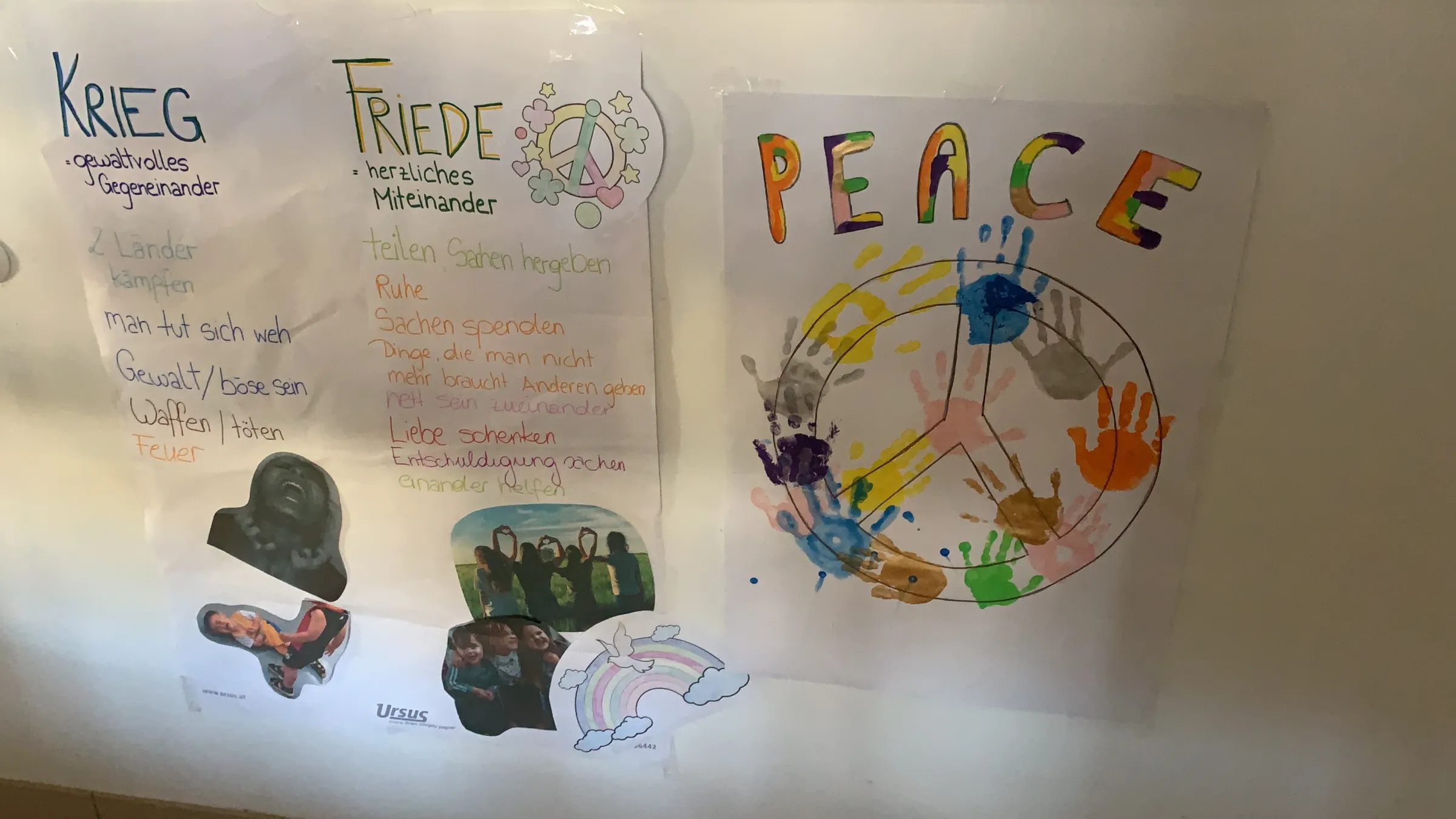 Drei Poster an einer Wand: links 'KRIEG' mit Stichpunkten, Mitte 'FRIEDE' mit Text, rechts farbiges 'PEACE' mit Weltkugel und Handabdrücken.