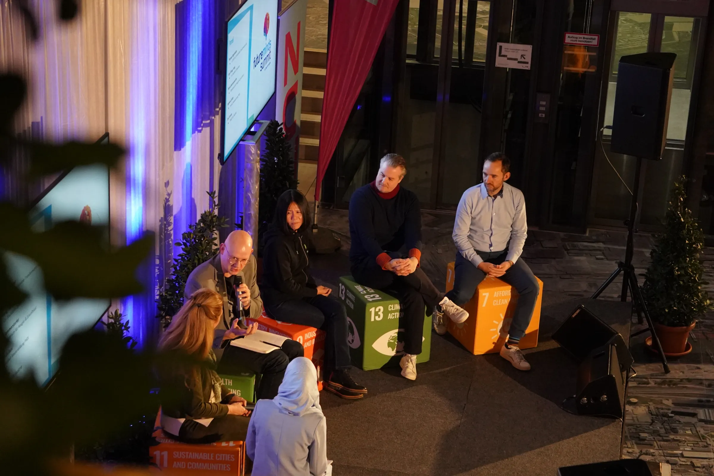 Paneldiskussion im Konferenzraum: Fachleute sitzen auf bunten Würfeln mit SDG-Zahlen, Moderator spricht ins Mikrofon.