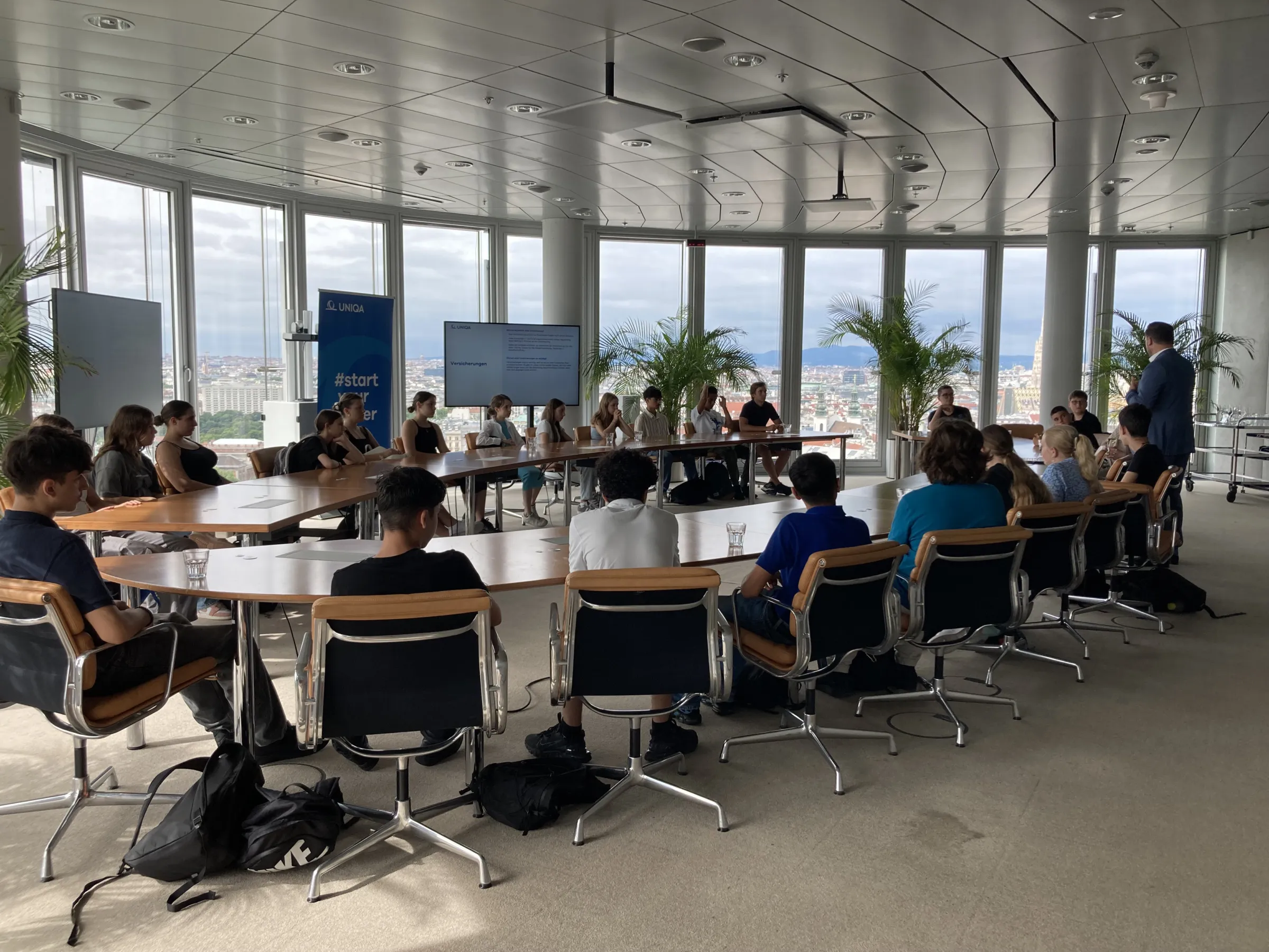 Seminarteilnehmende sitzen um einen großen Konferenztisch; rechts spricht eine Person; Fensterfront mit Stadtblick, Pflanzen im Raum.