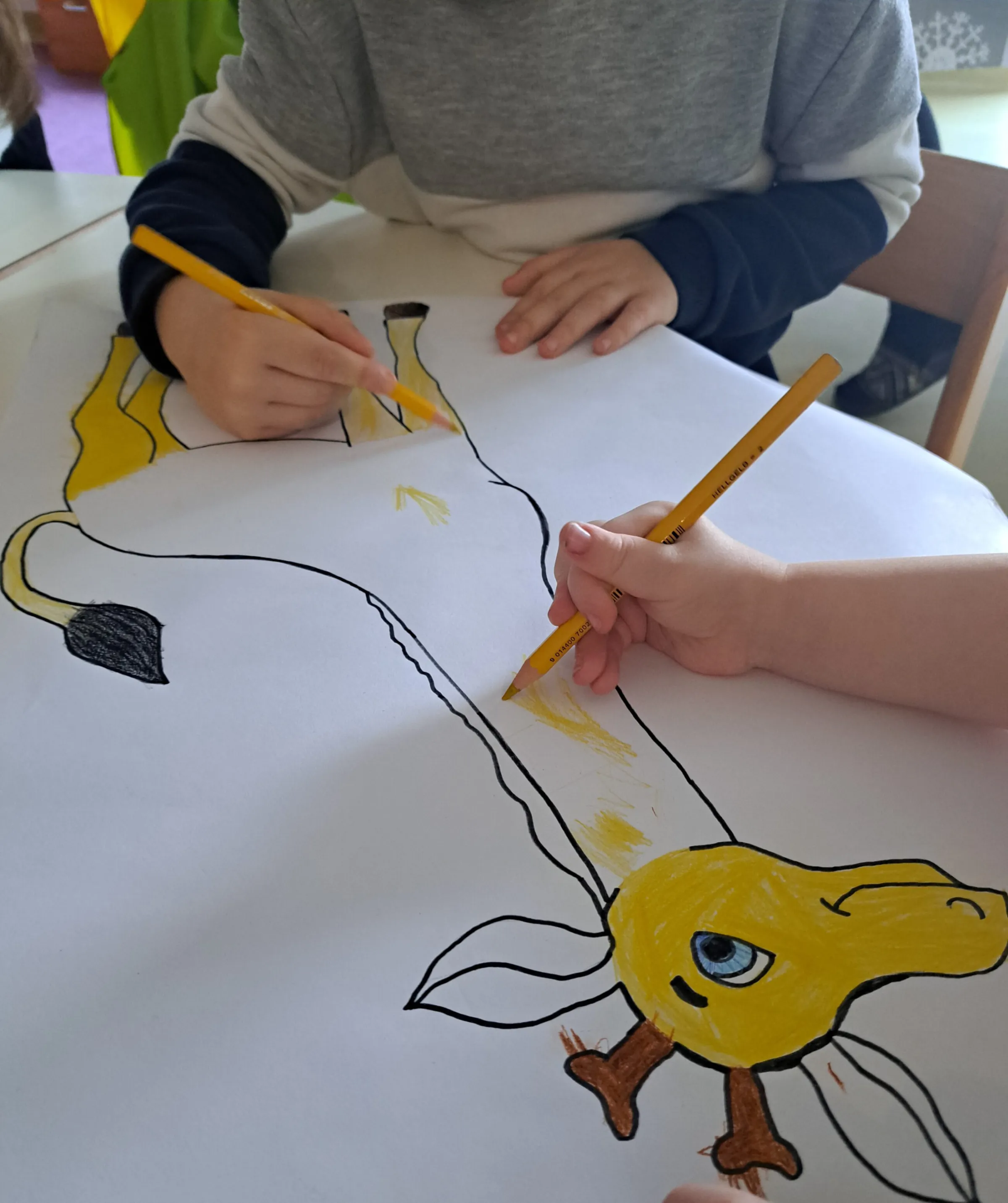 Zwei Kinder malen eine gelbe Giraffenzeichnung auf Papier am Tisch aus.