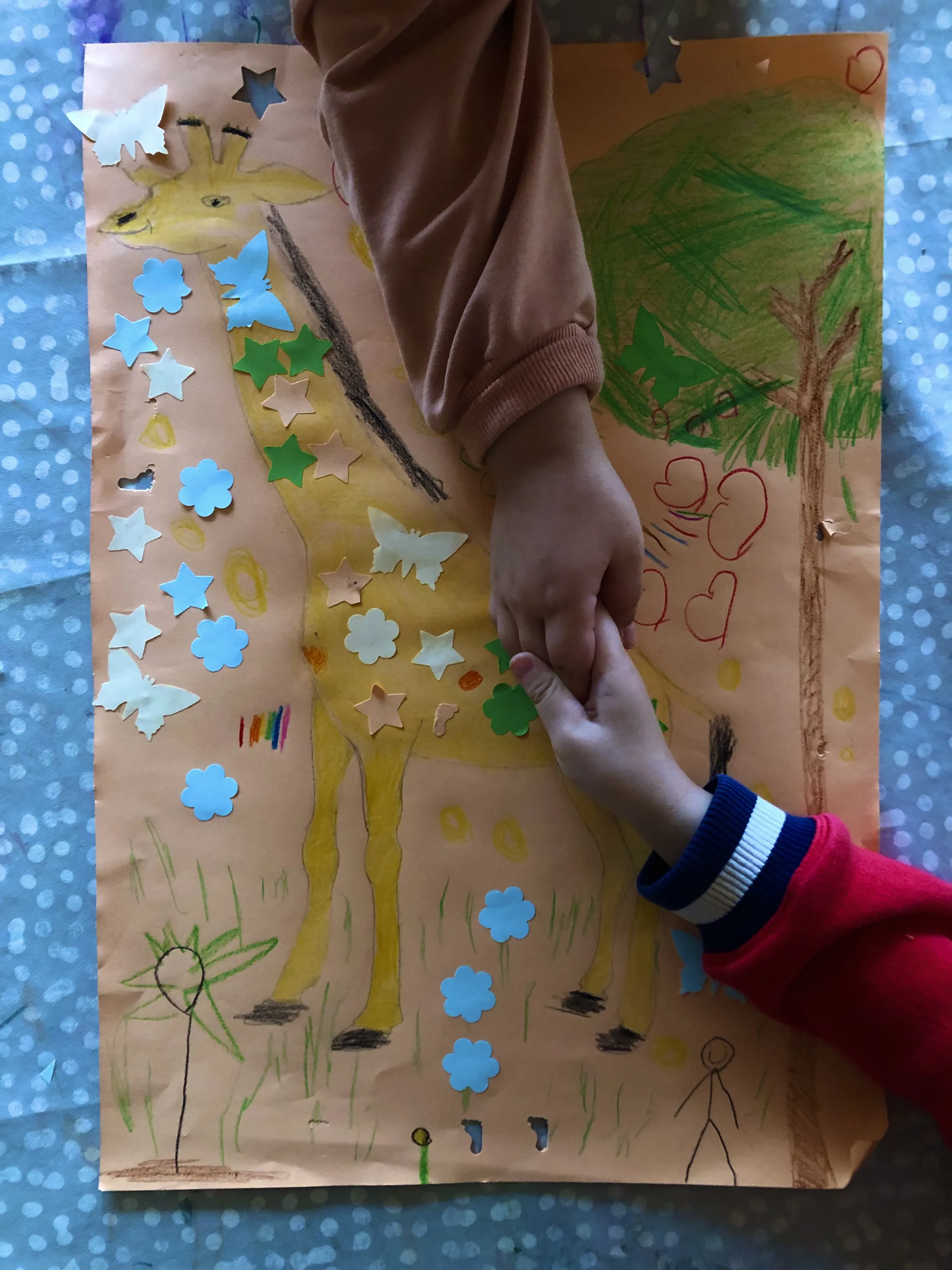 Zwei Kinderhände berühren sich über einem orangefarbenen Plakat mit gelber Giraffe, Bäumen und bunten Aufklebern vor blau gepunktetem Hintergrund.