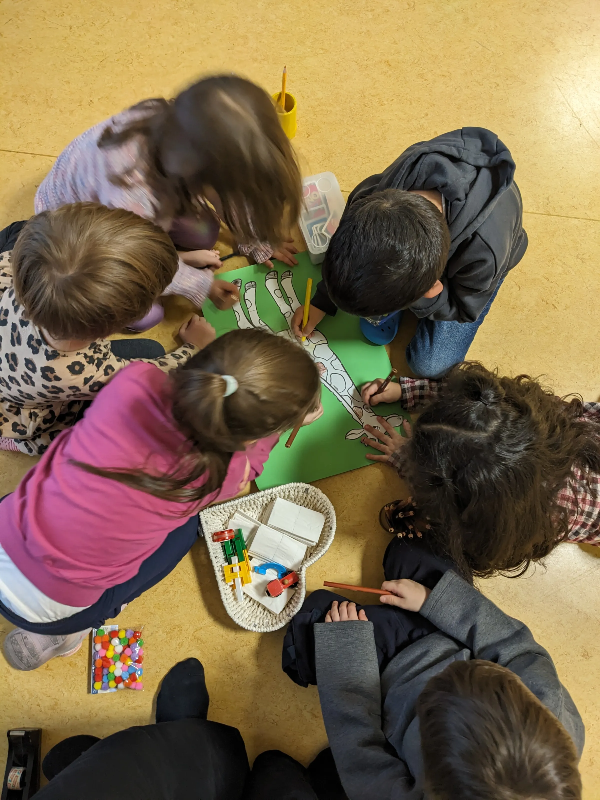 Kindergruppe sitzt auf dem Boden und bemalt sowie schneidet eine grüne Bastelvorlage, umgeben von Bastelmaterialien.