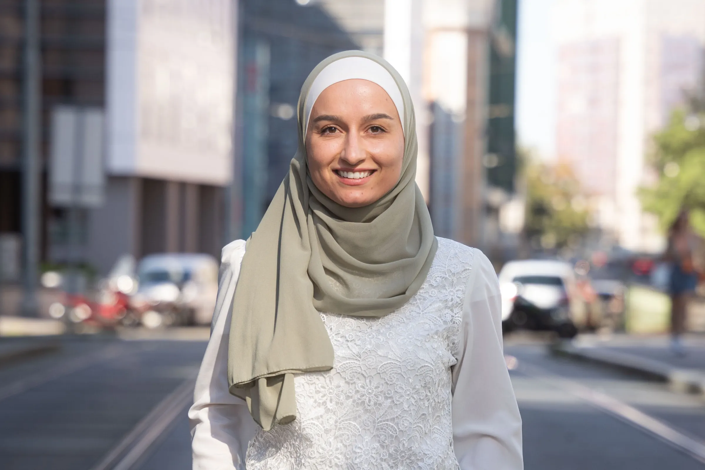Person mit hellgrünem Hijab und weißem Spitzenoberteil steht dort, lächelnd vor urbaner Straßenszene