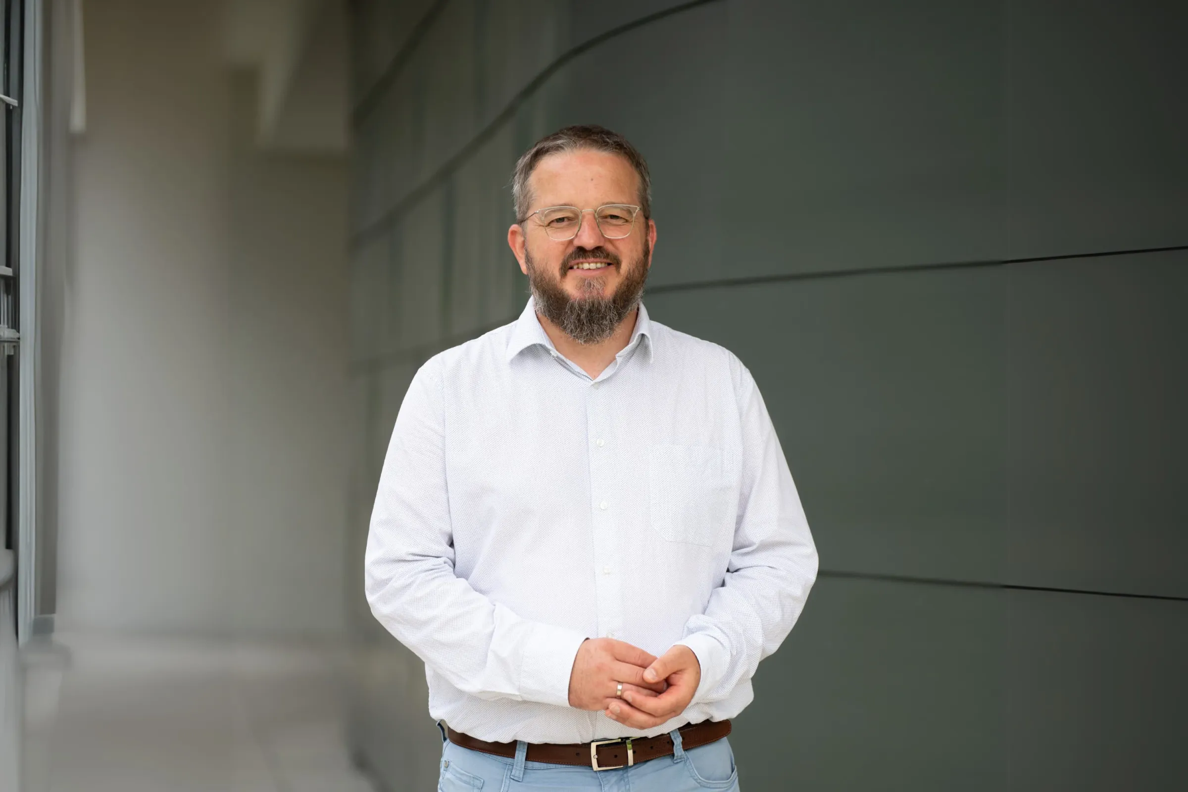 Person mit Bart und Brille, weißes Hemd, Jeans, brauner Gürtel, lächelnd, steht, Hände vor dem Bauch, vor grauer Wandverkleidung.