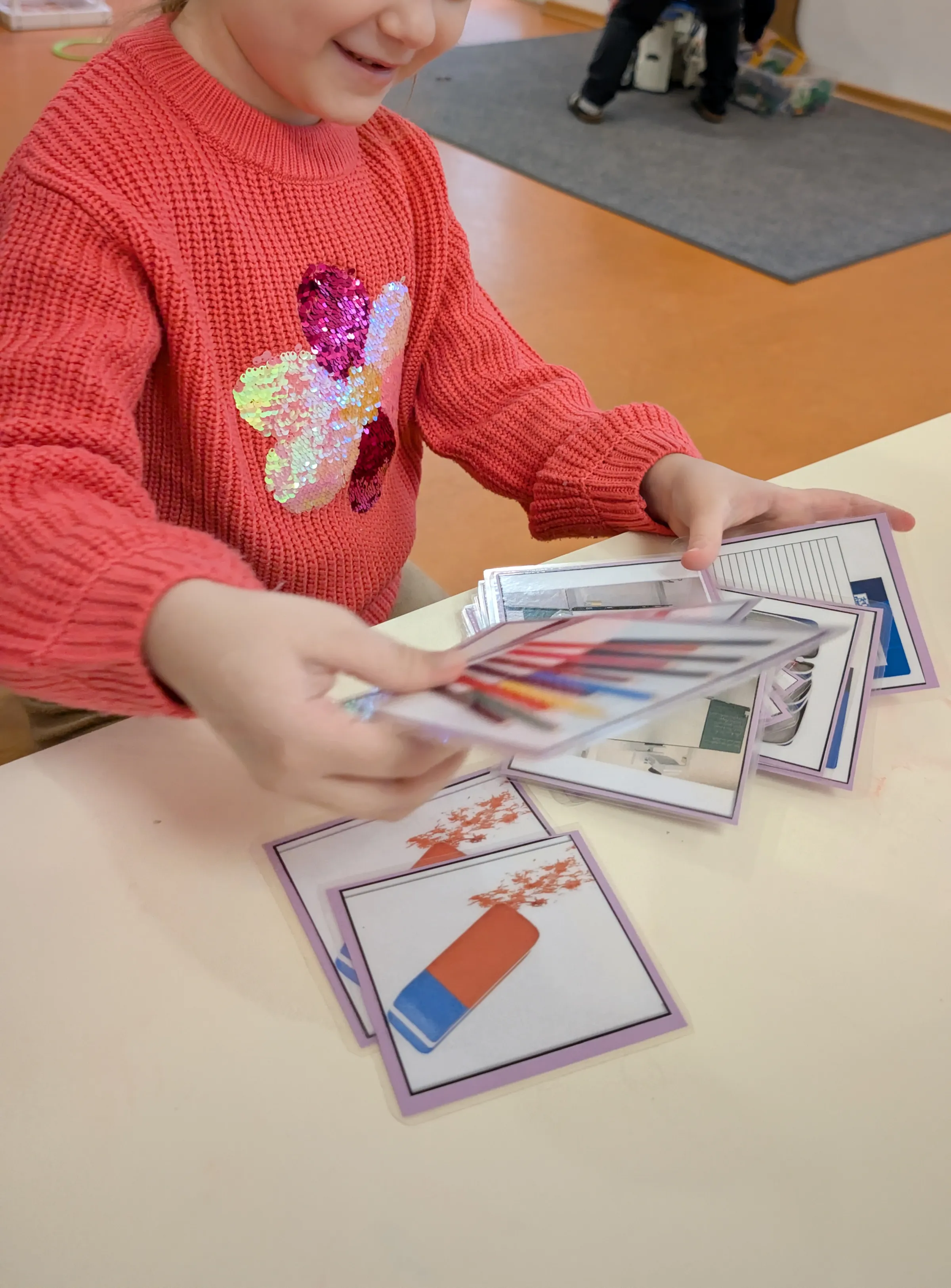Ein Kind in rotem Strickpullover mit einer glitzernden Blumenapplikation hält bunte Spielkarten auf einem Tisch; im Hintergrund Spielbereich.