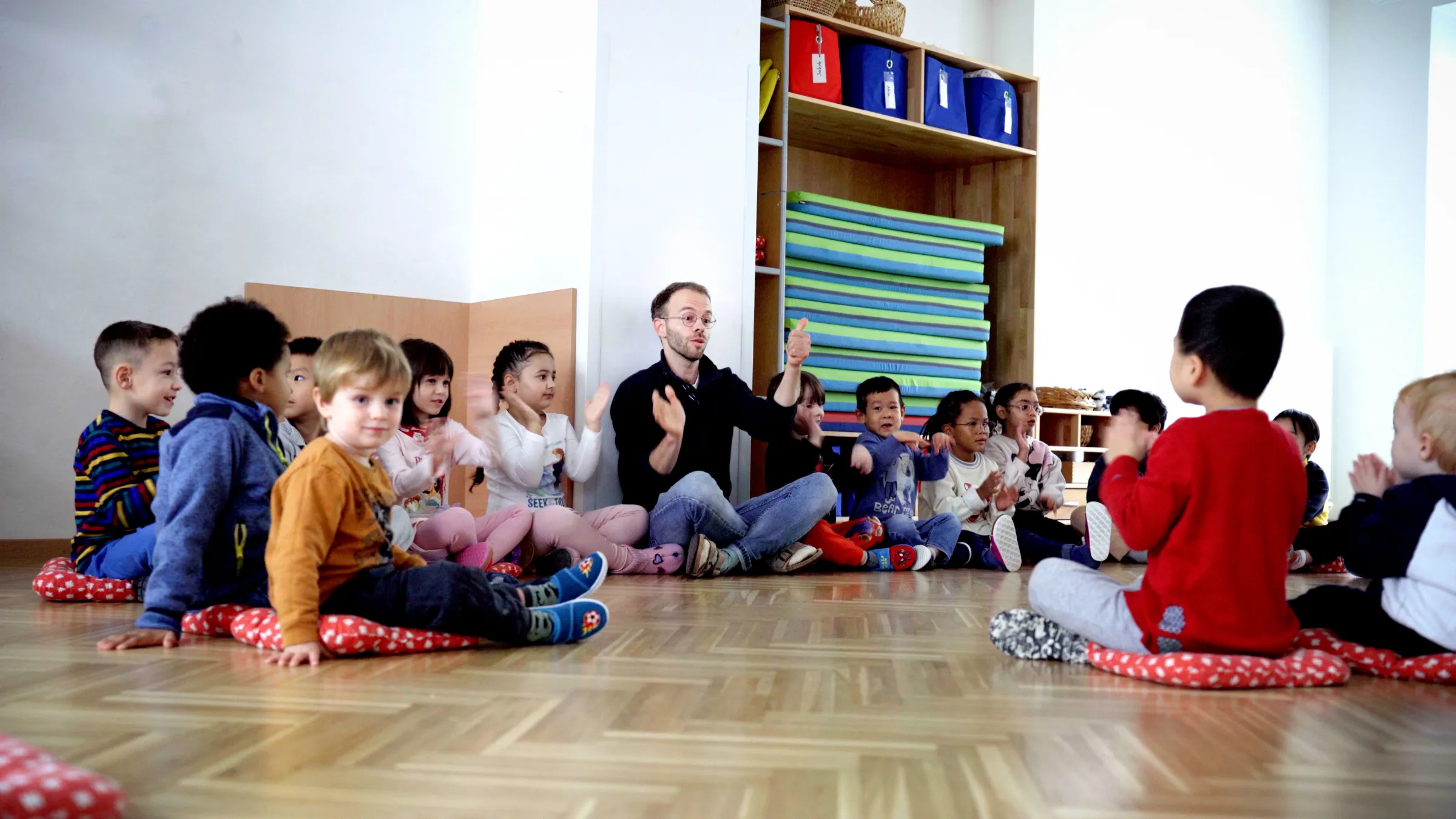Pädagogische Fachkraft sitzt in der Mitte eines Kreises von Kindern auf roten Sitzkissen; alle klatschen.