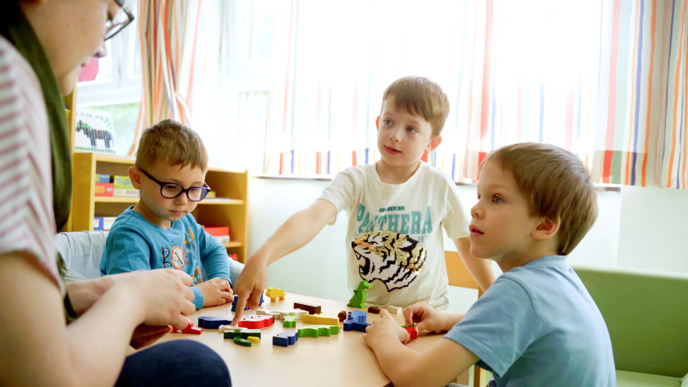 Vier Kinder im Klassenzimmer spielen mit bunten Bausteinen am Tisch; Lehrkraft schaut zu.