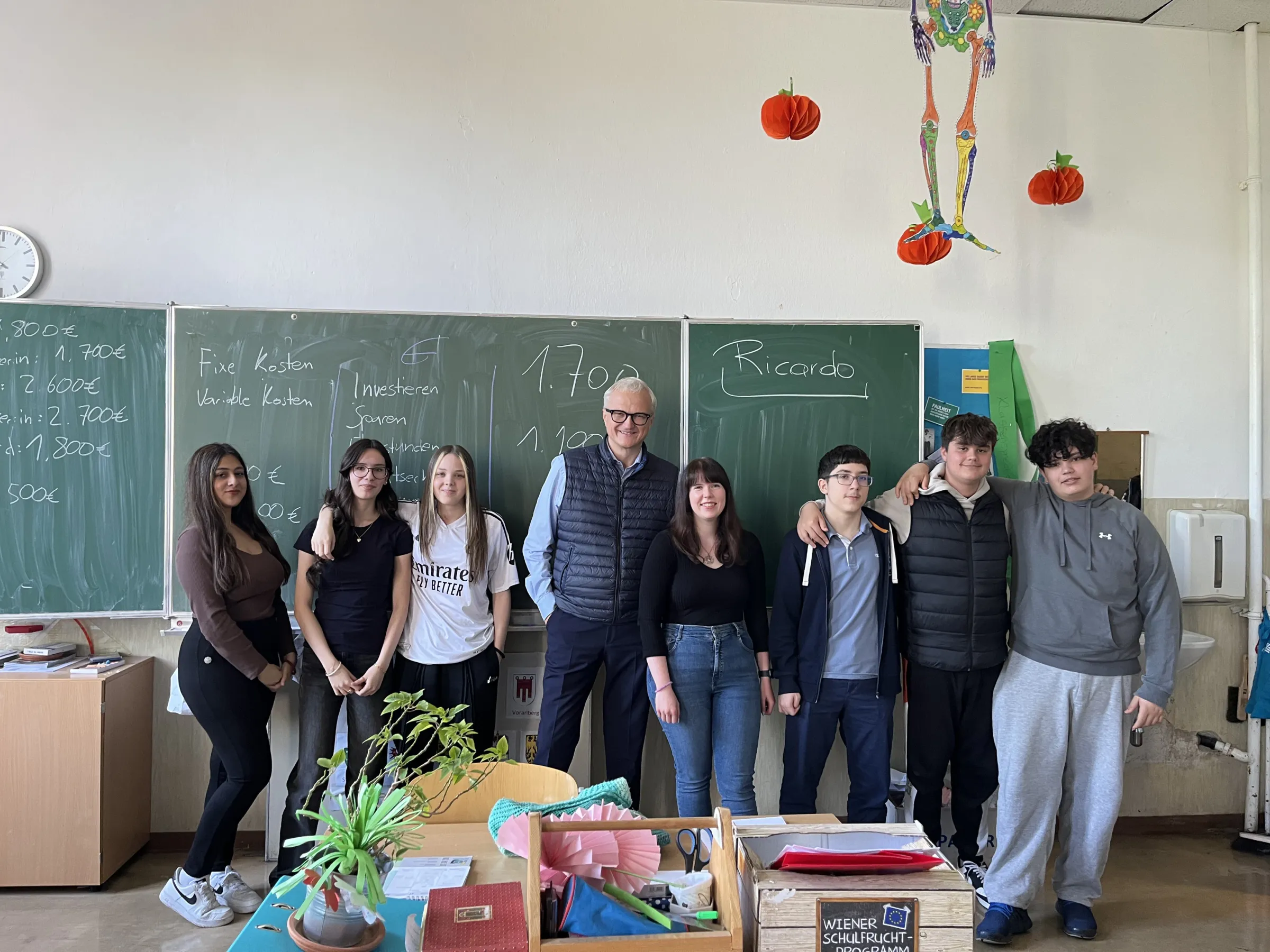 Gruppe aus acht Personen – Lehrkraft inklusive – posiert im Klassenzimmer vor grünen Tafeln; Kürbis-Dekorationen und Pflanzen auf dem Tisch.