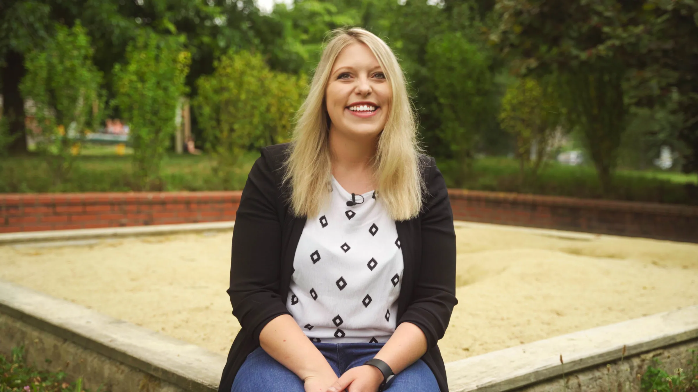 Blonde Person sitzt lächelnd im Park vor Sandkasten, trägt weiße Bluse mit Diamant-Muster und schwarze Jacke.
