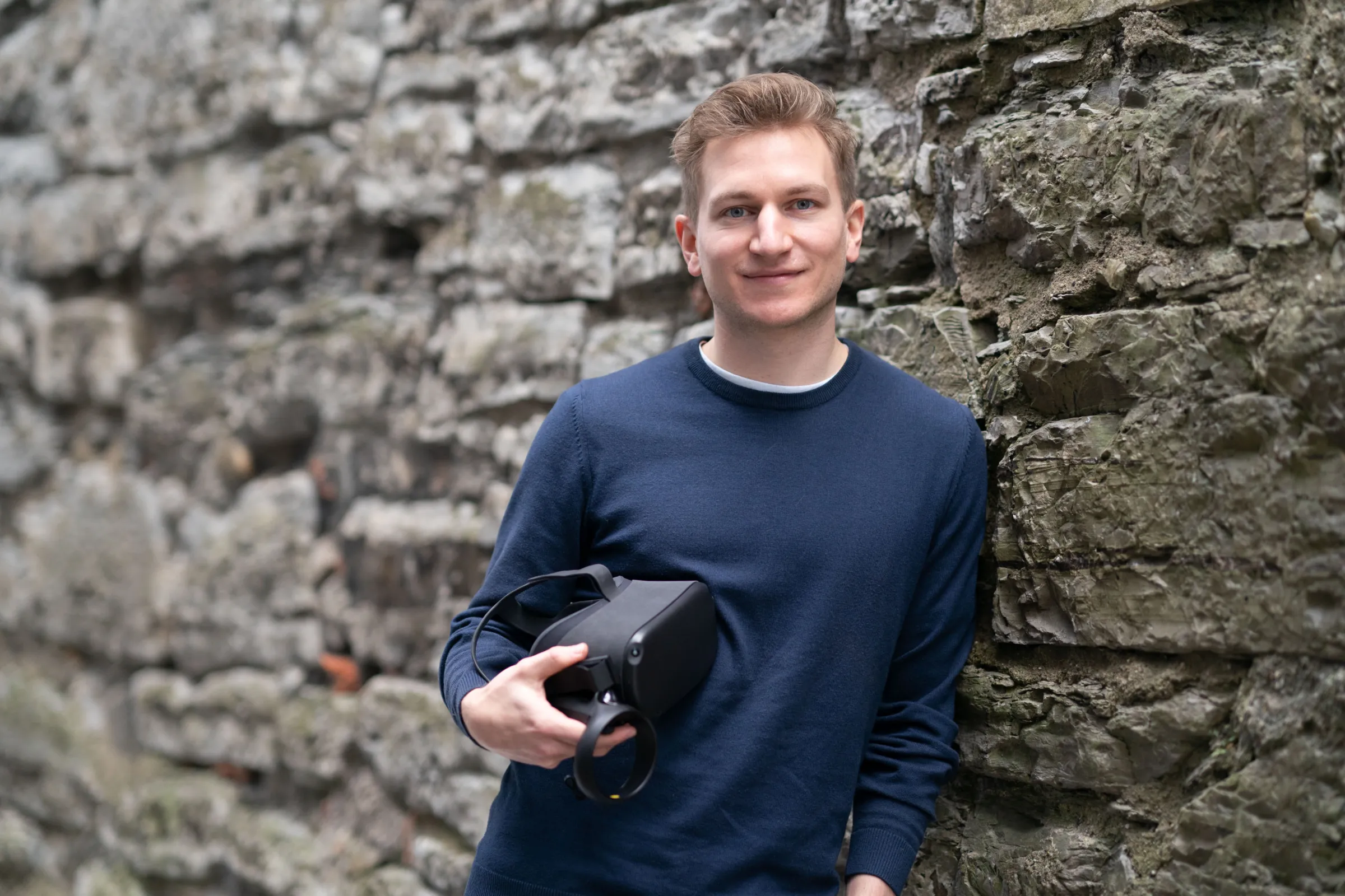Person lehnt an einer rauen Steinmauer und hält ein VR-Headset in der Hand