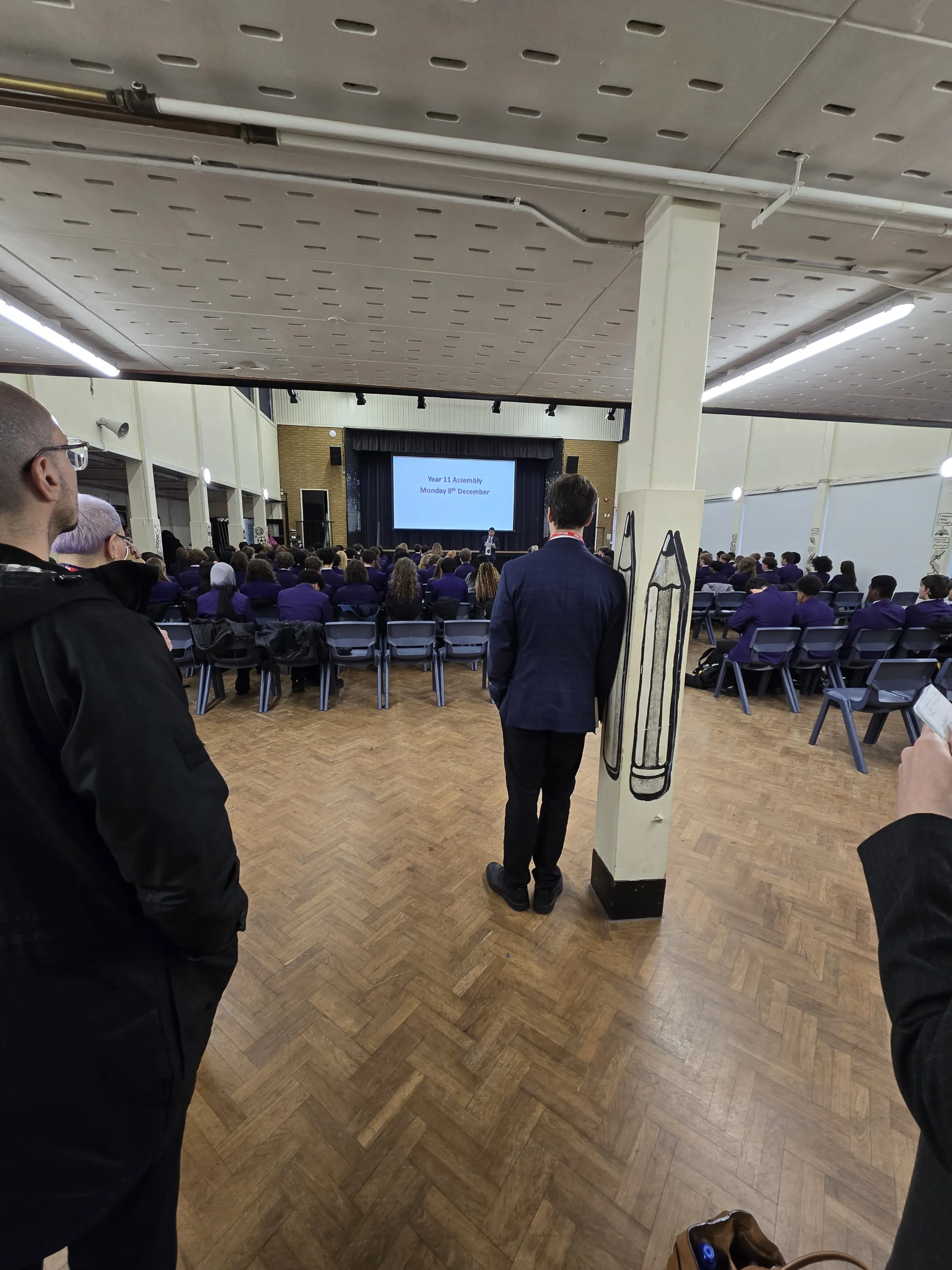 Großer Konferenzsaal mit Holzfußboden; viele Personen in lila Kleidung sitzen in Reihen, vorne Bühne mit Bildschirm.