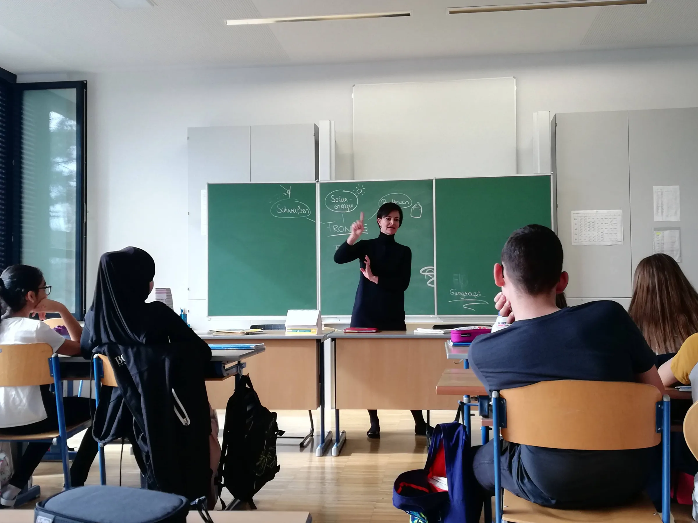 Lehrkraft erklärt Inhalte vor grüner Tafel im Klassenzimmer.