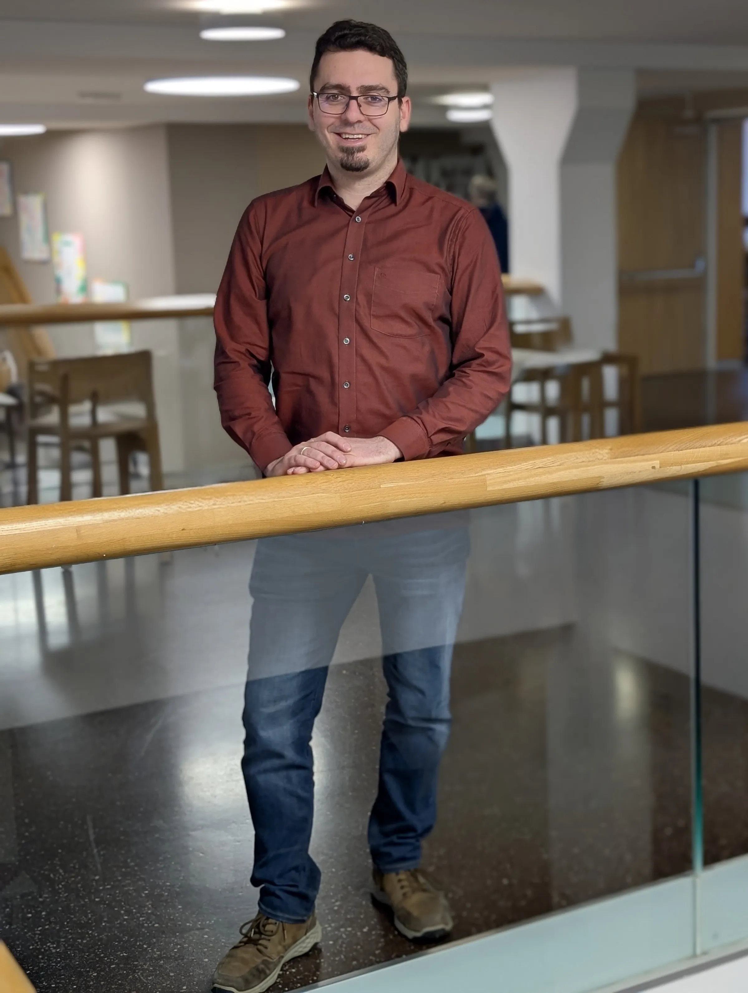 Person mit Brille steht lächelnd hinter Holzgeländer in modernem Innenraum, trägt dunkelrotes Hemd und Jeans.