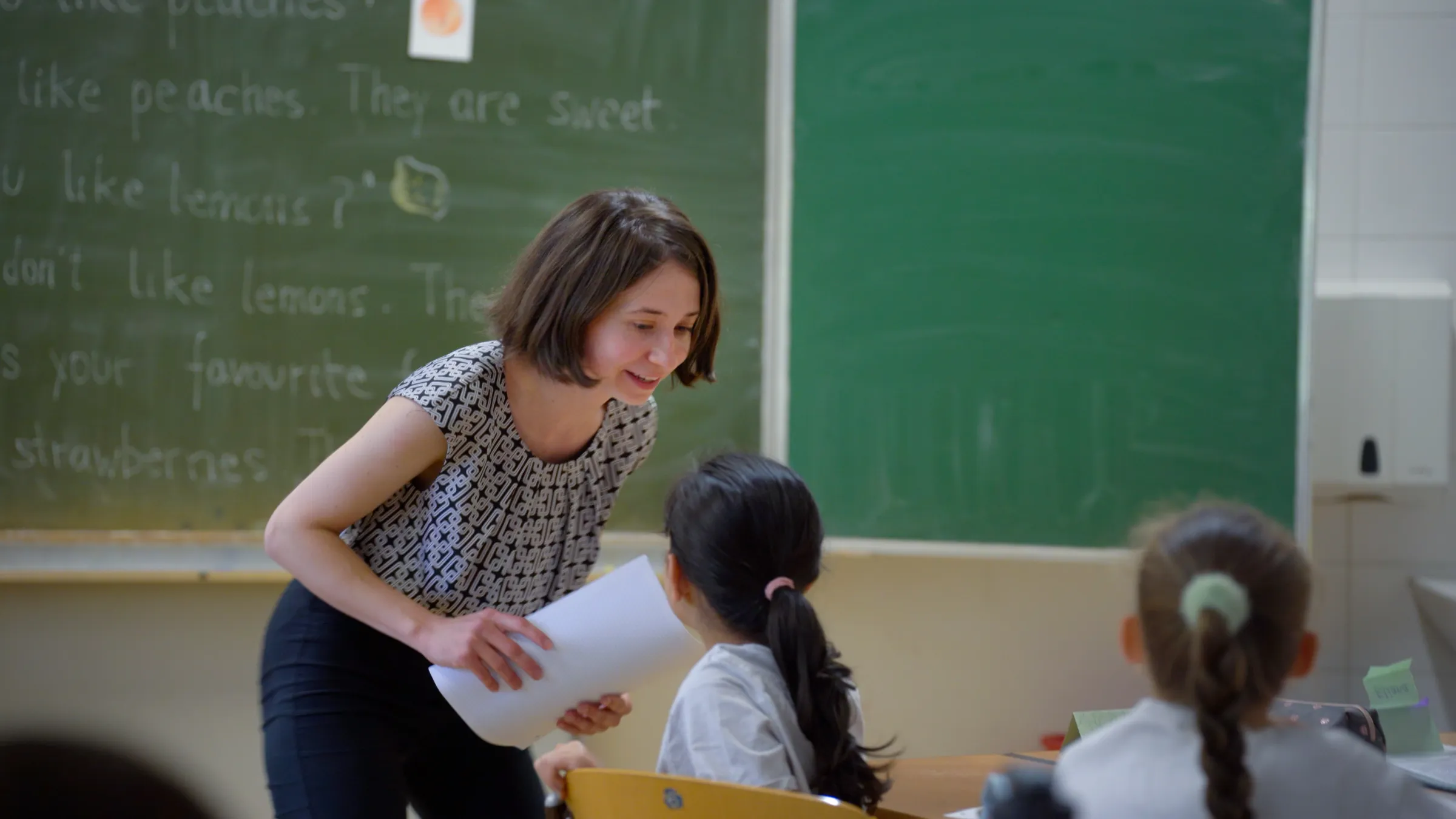 Lehrperson reicht einem Kind ein Blatt in einem Klassenraum; grüne Tafel im Hintergrund.