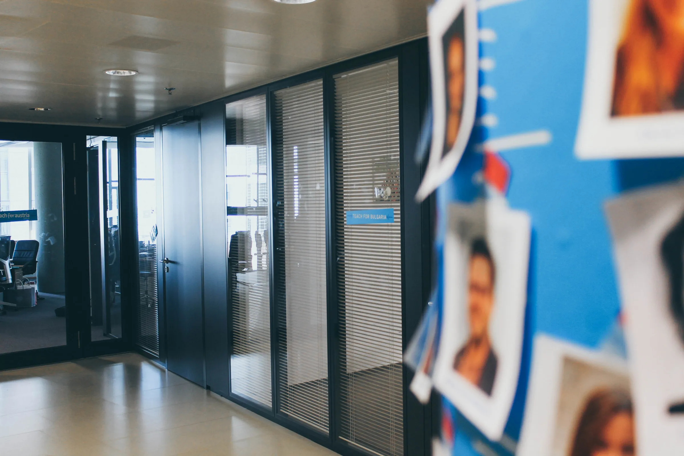 Leerer Büroflur mit Glastrennwänden und Jalousien; rechts bunte Posterwand mit Fotos.