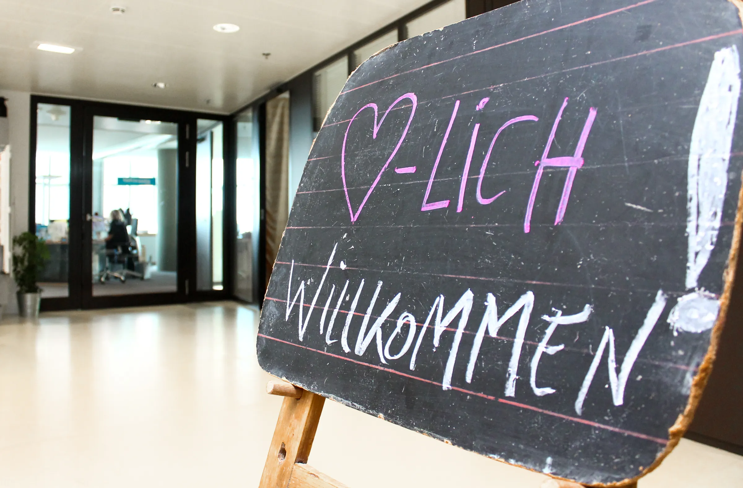 Schwarze Schiefertafel auf Holzständer im Empfangsbereich eines Büros; pinke Schrift "HERZLICH WILLKOMMEN" als Willkommensgruß.