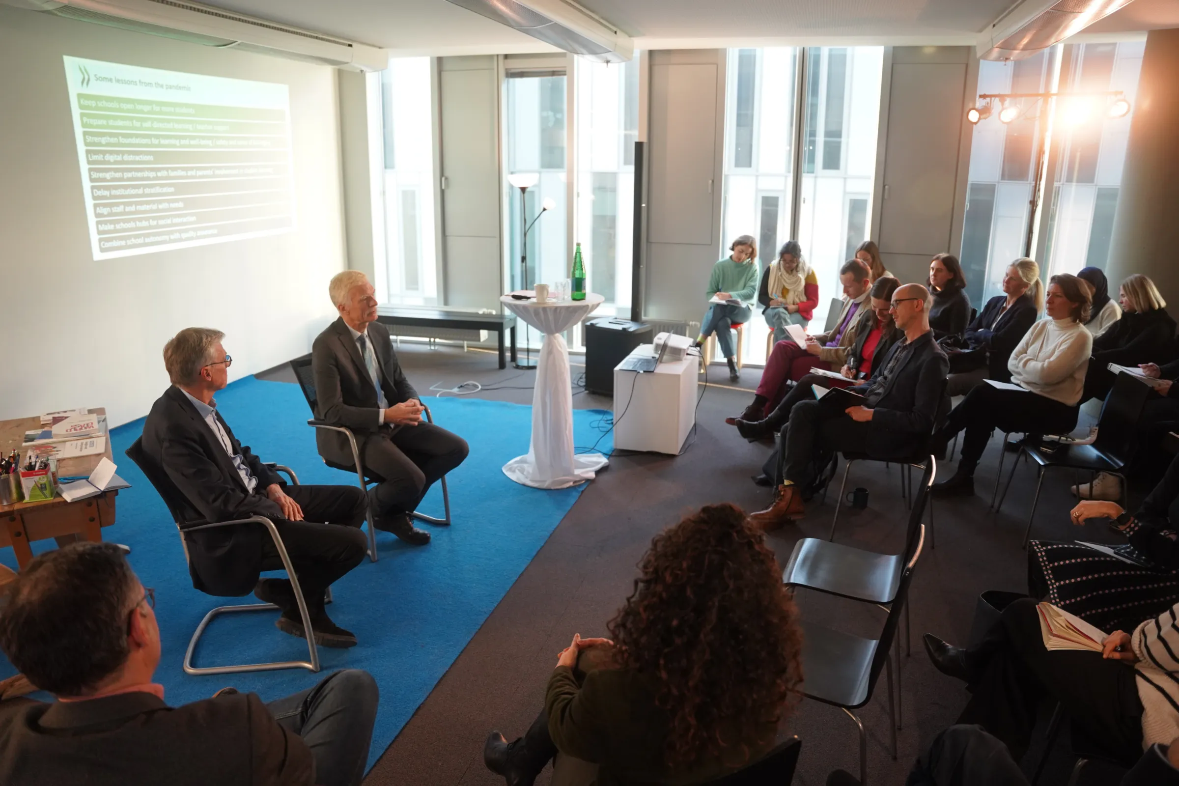 Zwei Redner im Anzug diskutieren vor Publikum in einem Konferenzraum; Beamer projiziert Folien, Zuhörer folgen der Präsentation.