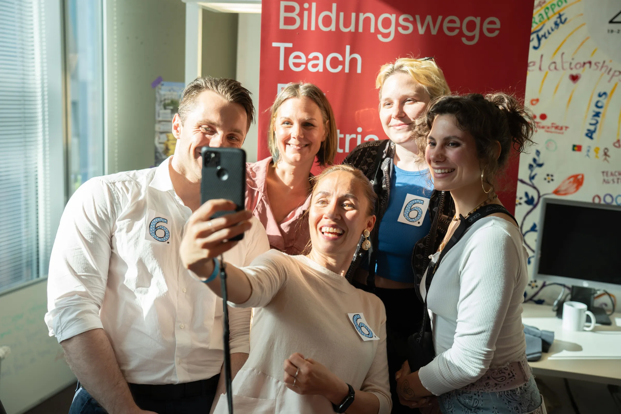 Gruppe von fünf Personen macht Selfie mit Smartphone; lächelt, nummerierte Abzeichen, im hellen Veranstaltungsraum.