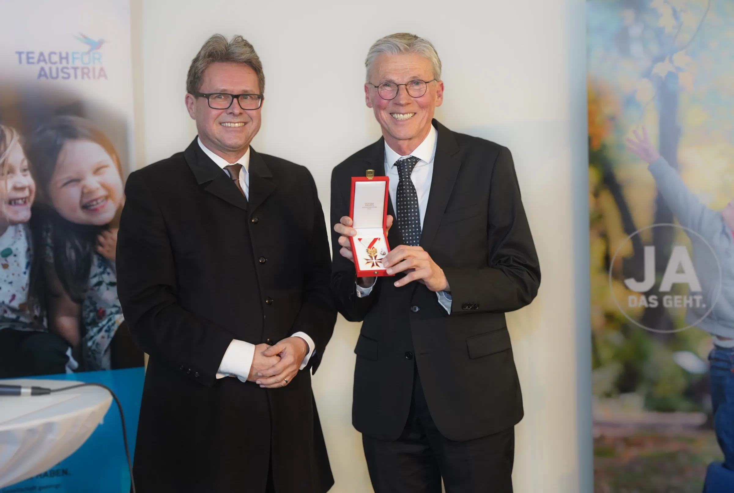 Zwei Männer in Anzügen; einer überreicht eine Medaille in roter Schatulle; im Hintergrund Banner.