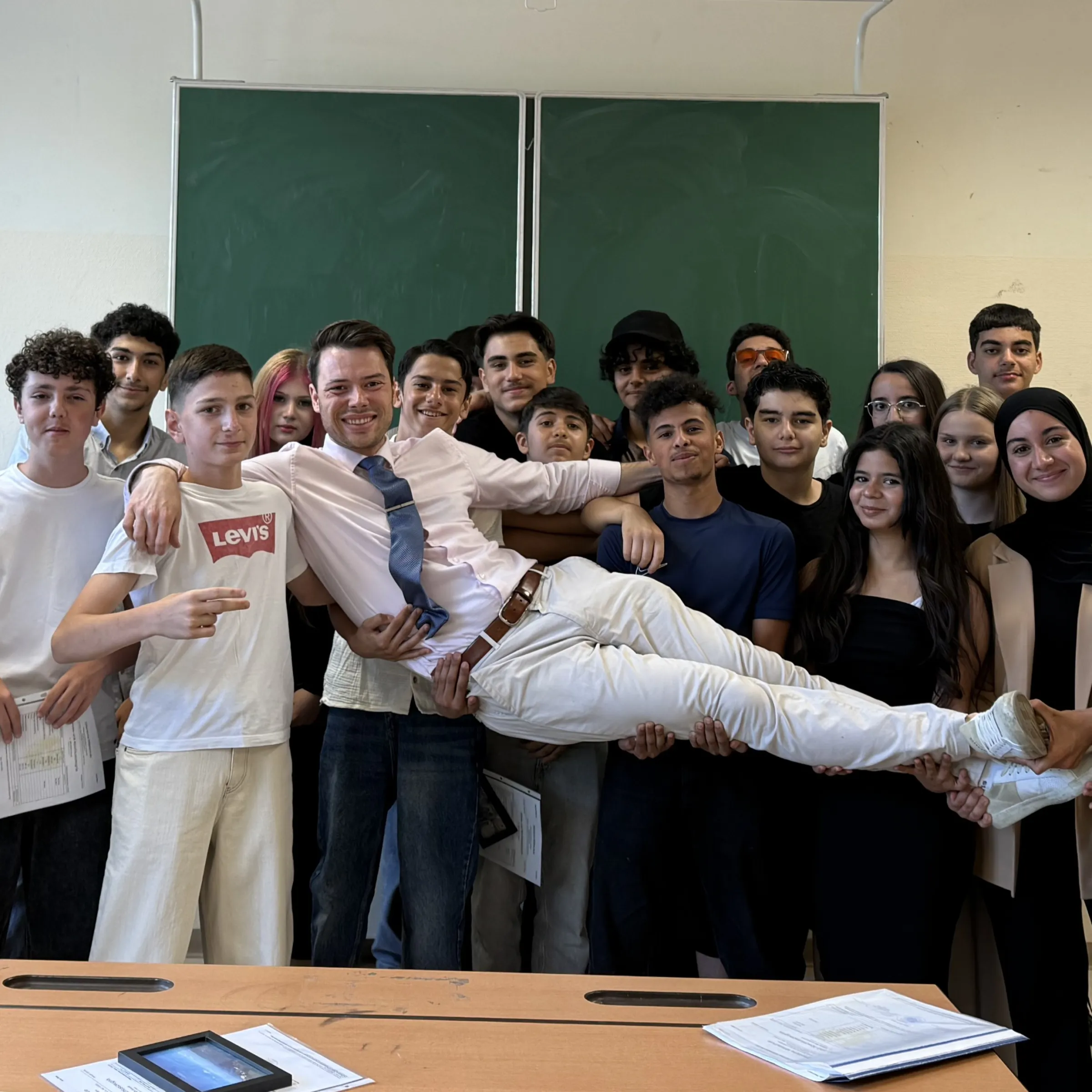 Lehrperson wird von einer Gruppe Lernender horizontal durchs Klassenzimmer getragen, vor grüner Tafel.