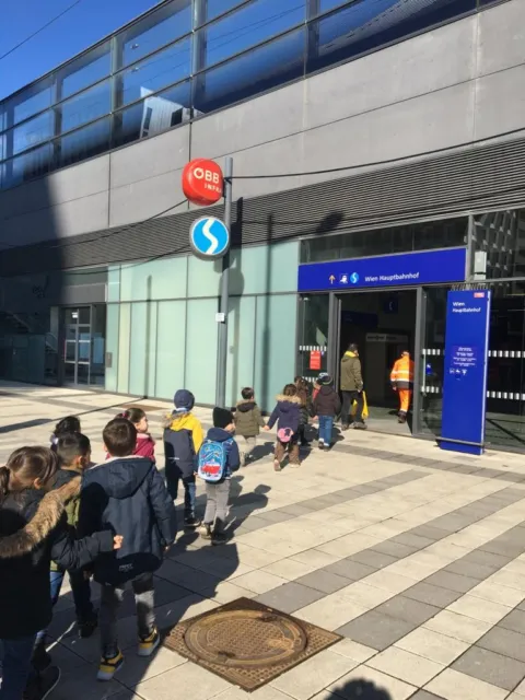 Gruppe Schulkinder mit Rucksäcken geht in einer Reihe zum Eingang des Wiener Hauptbahnhofs; modernes Glasgebäude, blaue Beschilderung.