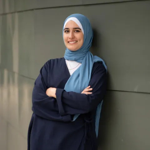 Person mit blauem Hijab lehnt an grüner Wand, verschränkte Arme, lächelt in die Kamera.