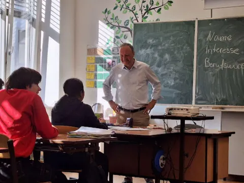 Lehrer steht vor zwei Schülern in einem Klassenzimmer und erklärt am Schreibtisch; Tafel im Hintergrund.