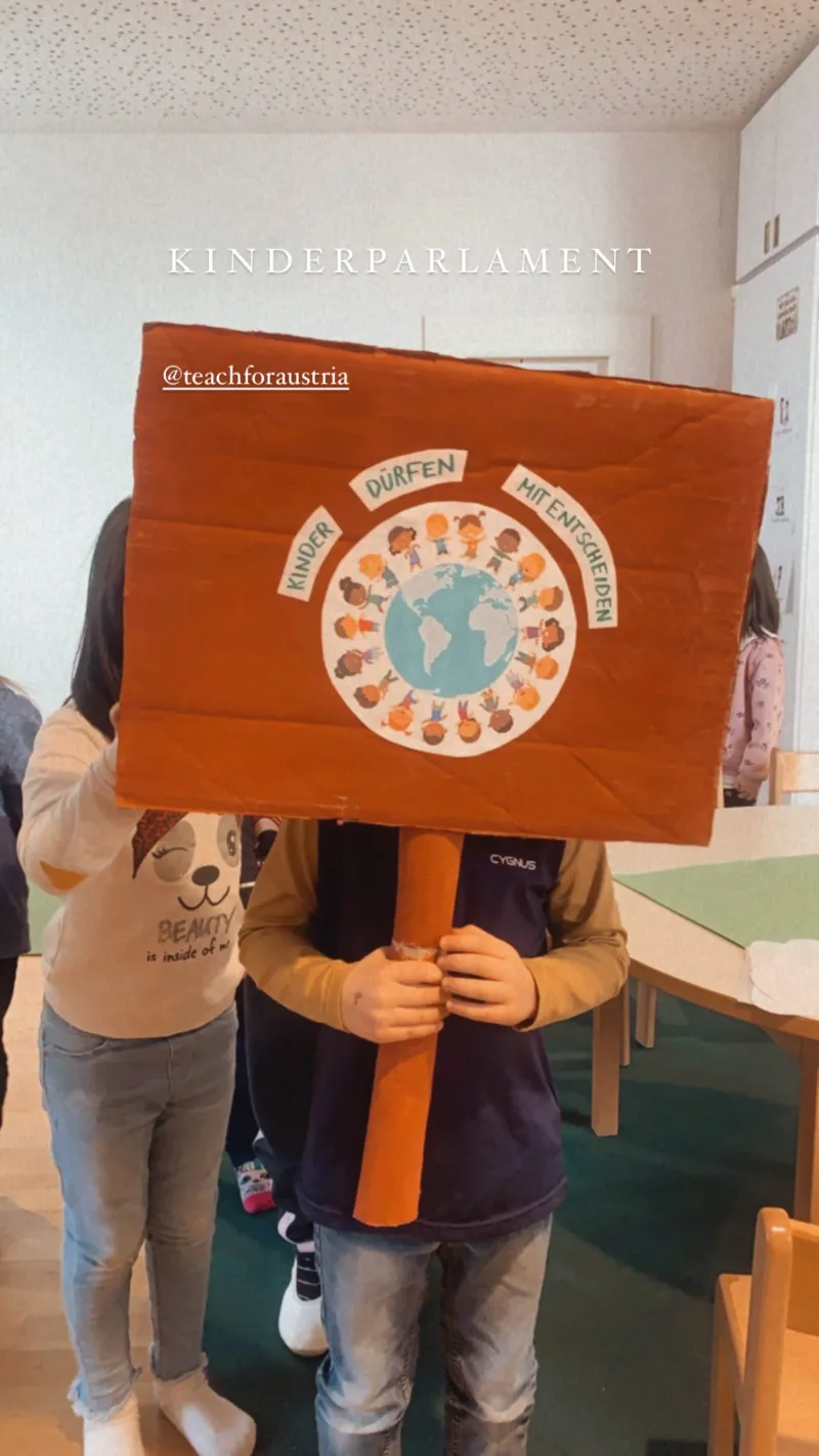 Ein Kind hält ein großes oranges Schild mit Globus und Kindern rundherum; Schulraum im Hintergrund.