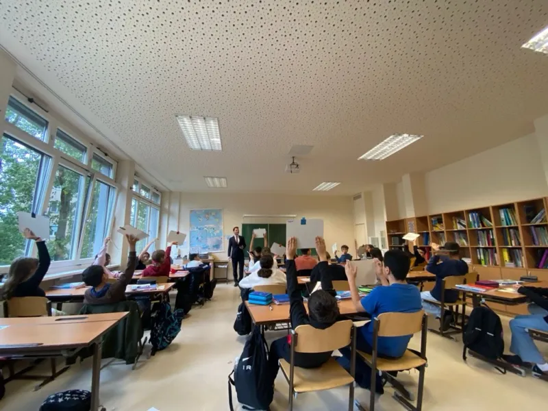 Klassenraum; Lehrperson steht vorne, Schüler heben weiße Zettel hoch; Bücherregale und Weltkarte an der Wand.