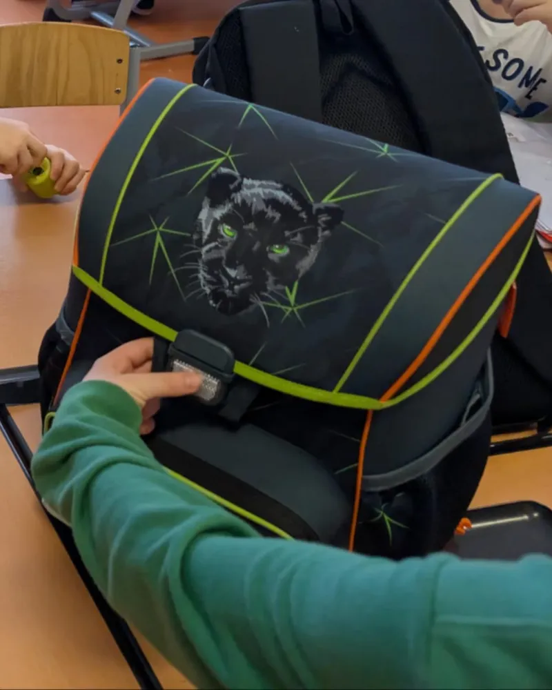 Schwarzer Schulrucksack mit grüner Panther-Grafik auf der Klappe; eine Person schließt die Schnalle im Klassenzimmer.