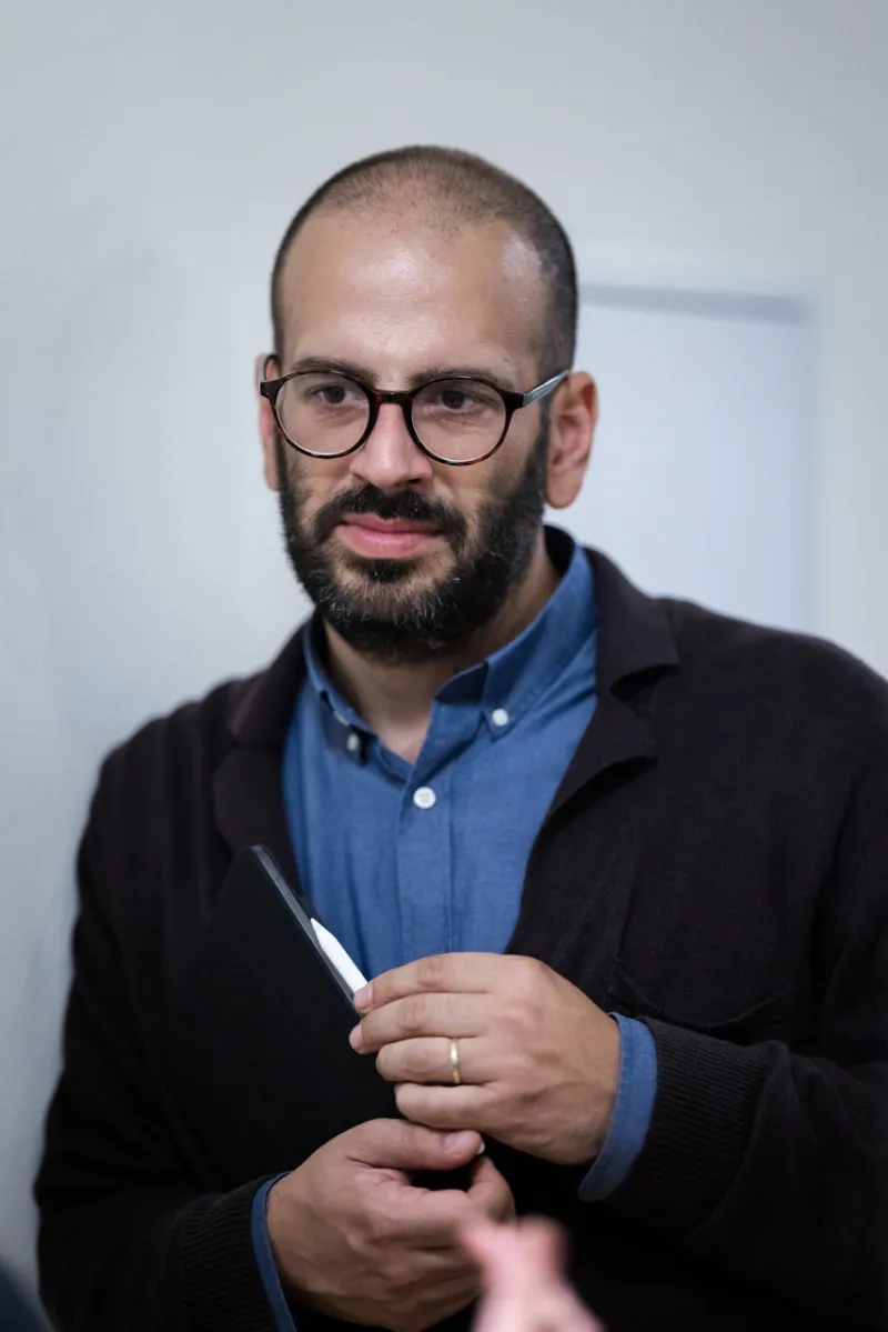Person mit runder Brille und Bart hält schwarzen Notizblock, trägt blauen Hemd und dunklen Cardigan, im Innenraum