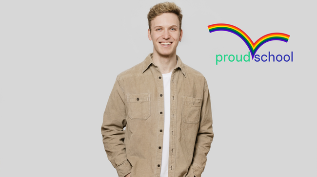 Junger Mann in beiger Jacke lächelt; Studiohintergrund mit Regenbogenlogo "proud school".