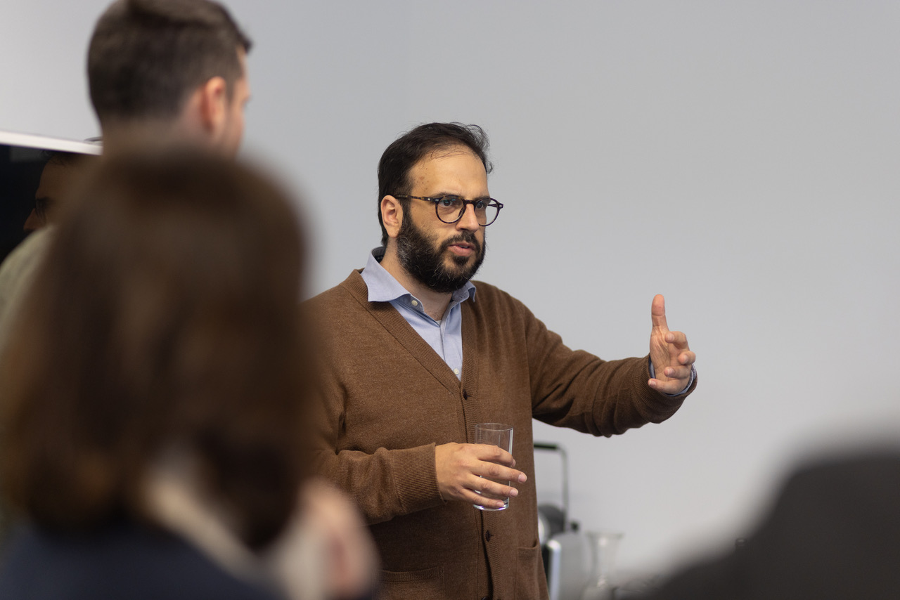 Referent mit Brille und Bart, brauner Cardigan, hält Glas und gestikuliert mit erhobenem Zeigefinger vor Publikum im Besprechungsraum.